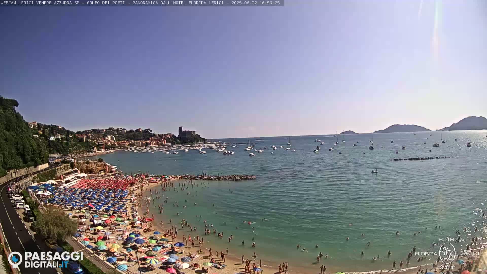 A crowded beach with colorful umbrellas under a clear blue sky, overlooks a calm bay filled with boats and distant hills.