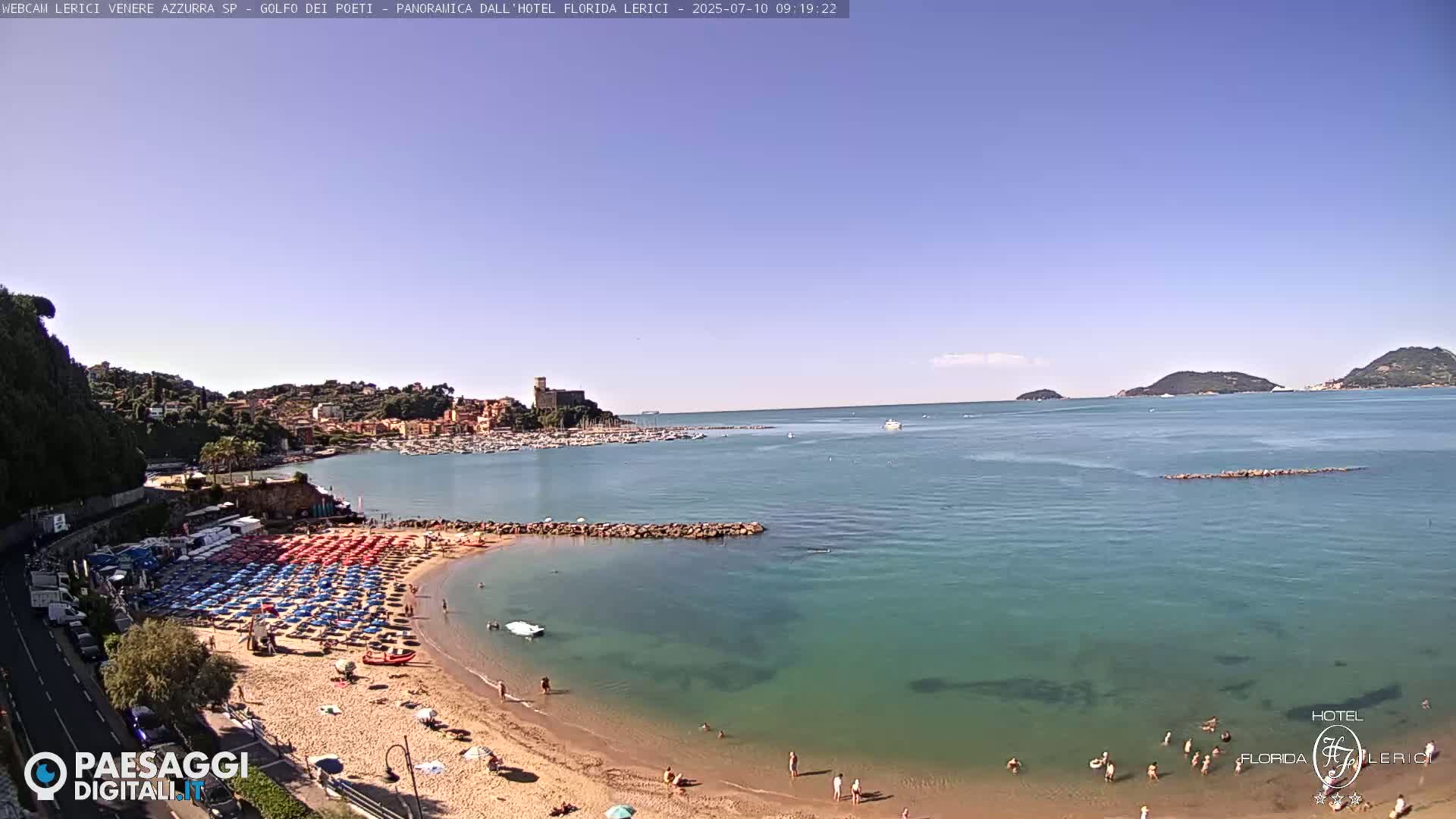 A sunny day shows a beach with many red and blue umbrellas, a calm teal sea, and a town with a castle in the distance.