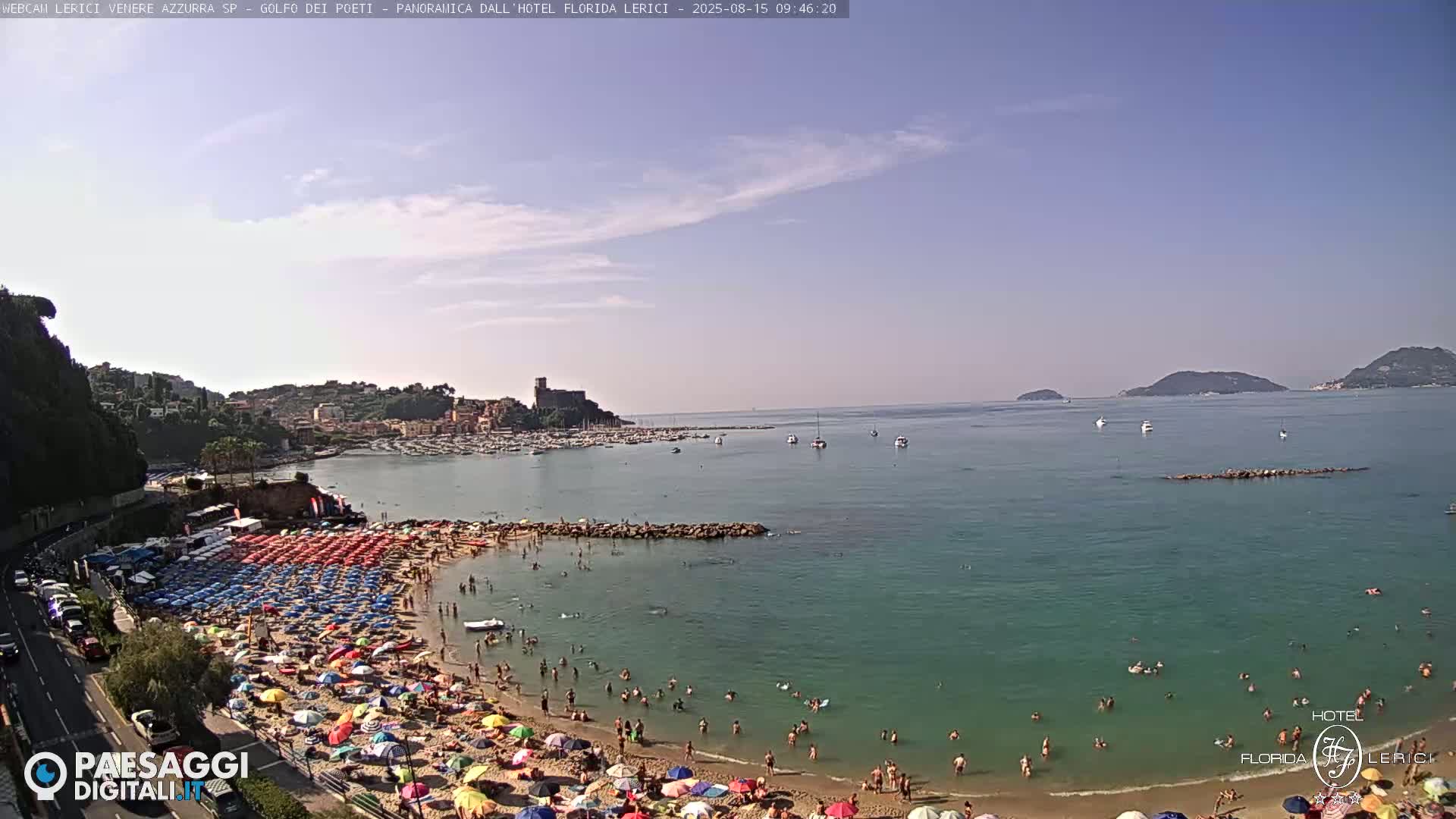 A crowded beach with colorful umbrellas under a mostly clear sky, overlooking a calm, teal sea with boats and islands in the distance.