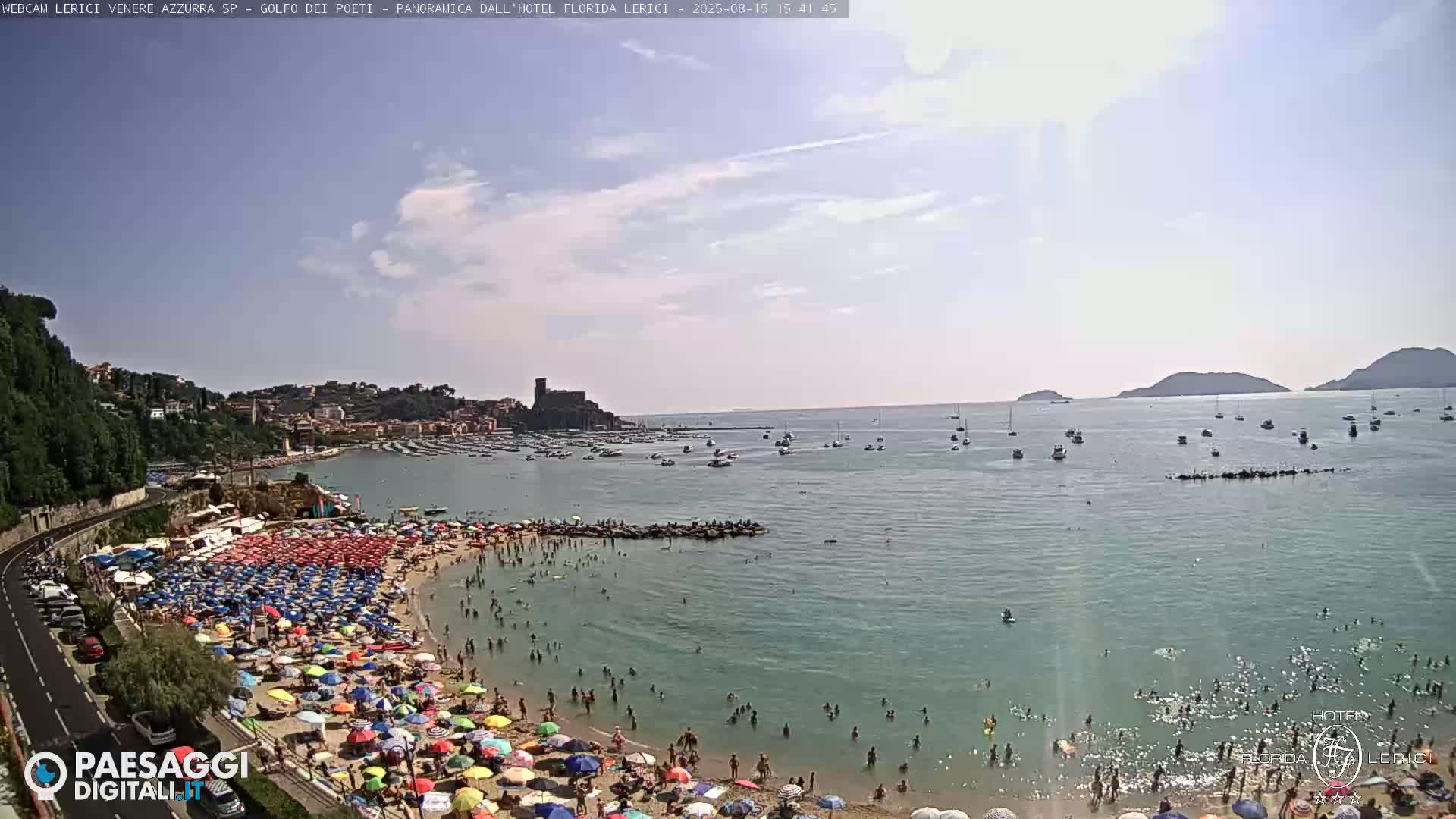A crowded beach with colorful umbrellas under a mostly clear sky, overlooking a calm, teal sea with boats and islands in the distance.
