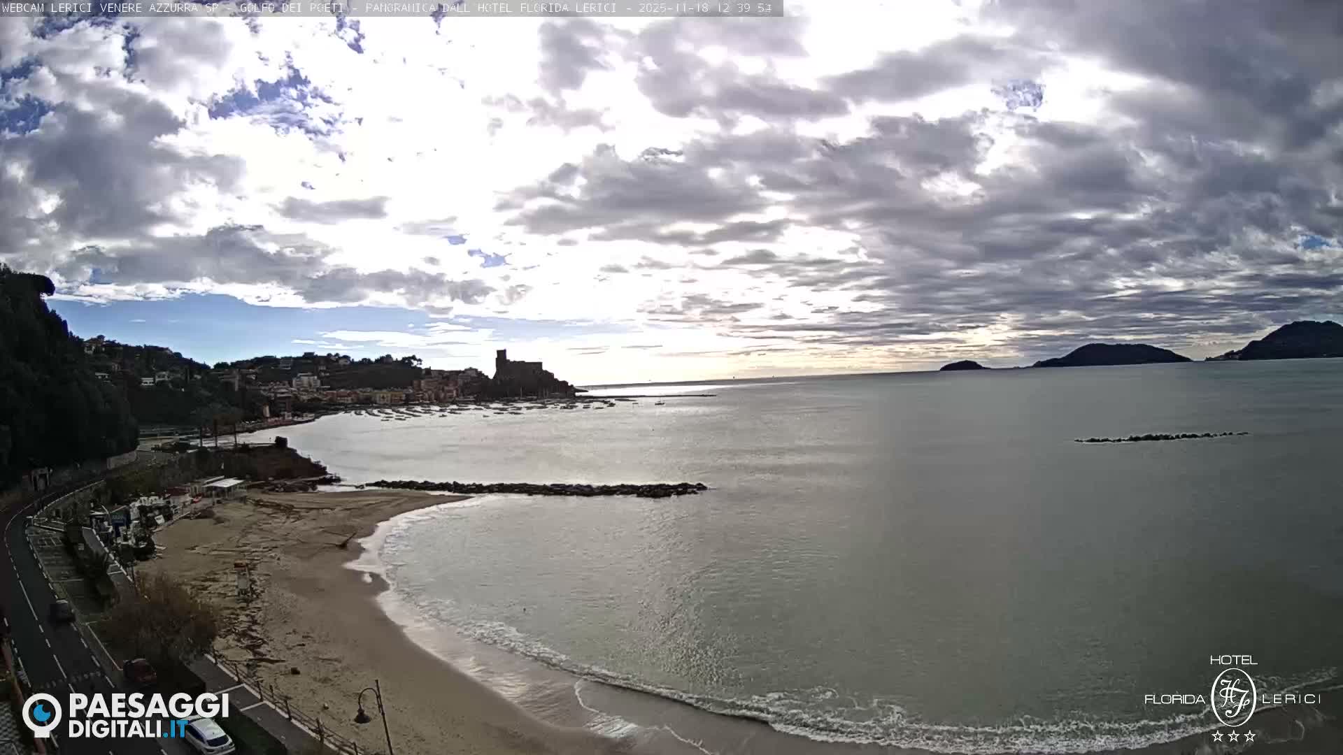 A clear night reveals a brightly lit coastal town and a prominent castle across a calm bay with gentle waves, bordered by a winding shoreline road.