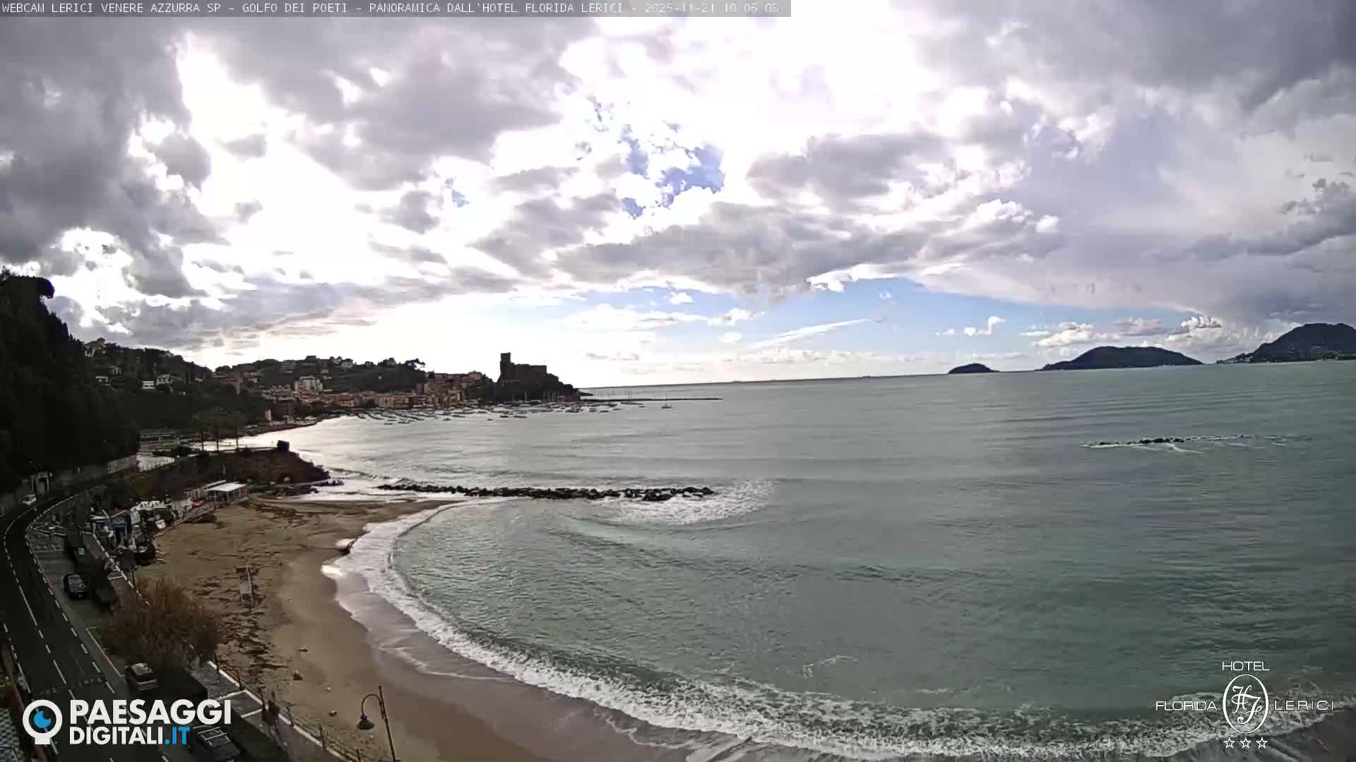 A panoramic coastal view captures a winding road alongside a sandy beach with gentle waves, leading to a town with a prominent castle on a tree-lined hillside, all overlooking a calm sea with distant islands under a bright, partly cloudy sky.
