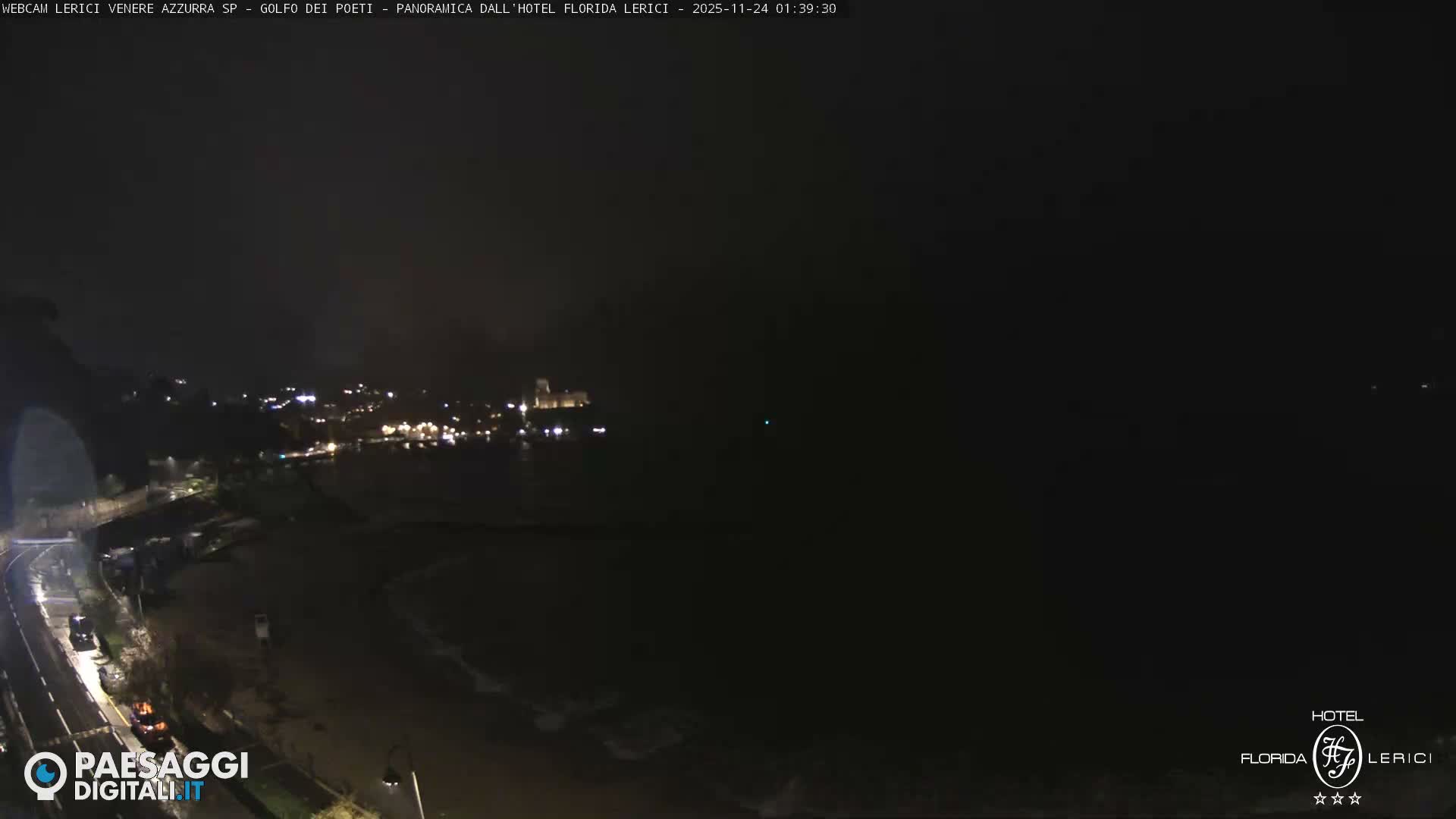 The image captures a dark, cloudy night scene of a coastal town with numerous illuminated buildings along a road and beach, facing a calm sea.