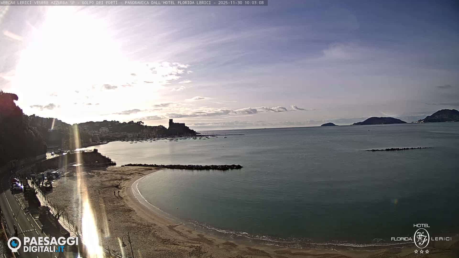 A panoramic view reveals a sunny coastal town featuring a sandy beach, a bay dotted with numerous boats, and a prominent castle, all under a bright, partly cloudy sky, with distant islands visible across the water.