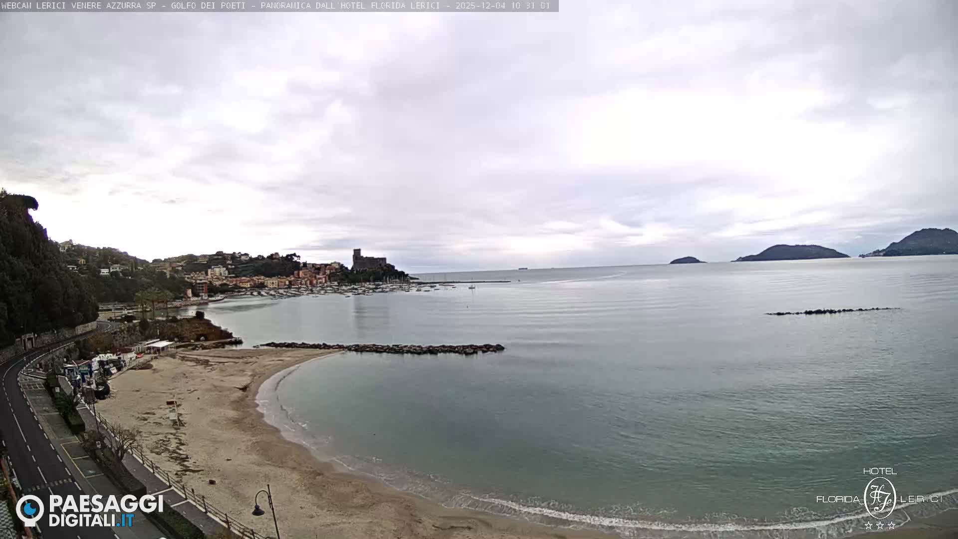 Under an overcast sky, a wide view captures a coastal town with a sandy beach, a harbor, a hilltop castle, and a calm sea dotted with distant islands.