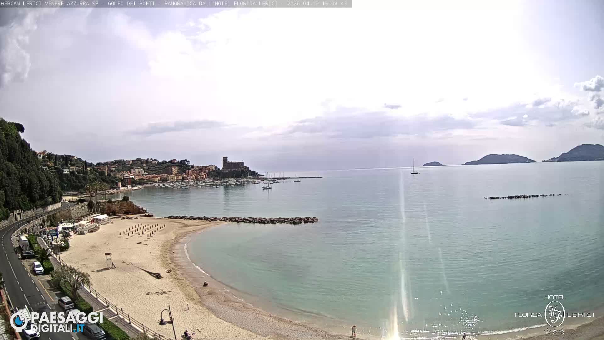 Under an overcast sky, a wide view captures a coastal town with a sandy beach, a harbor, a hilltop castle, and a calm sea dotted with distant islands.