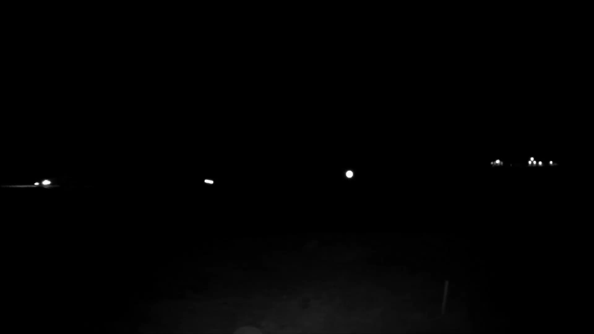 A clear, dark night scene features several distant, scattered white lights, likely from vehicles or structures, against an almost entirely black landscape with faint ground details visible at the bottom.