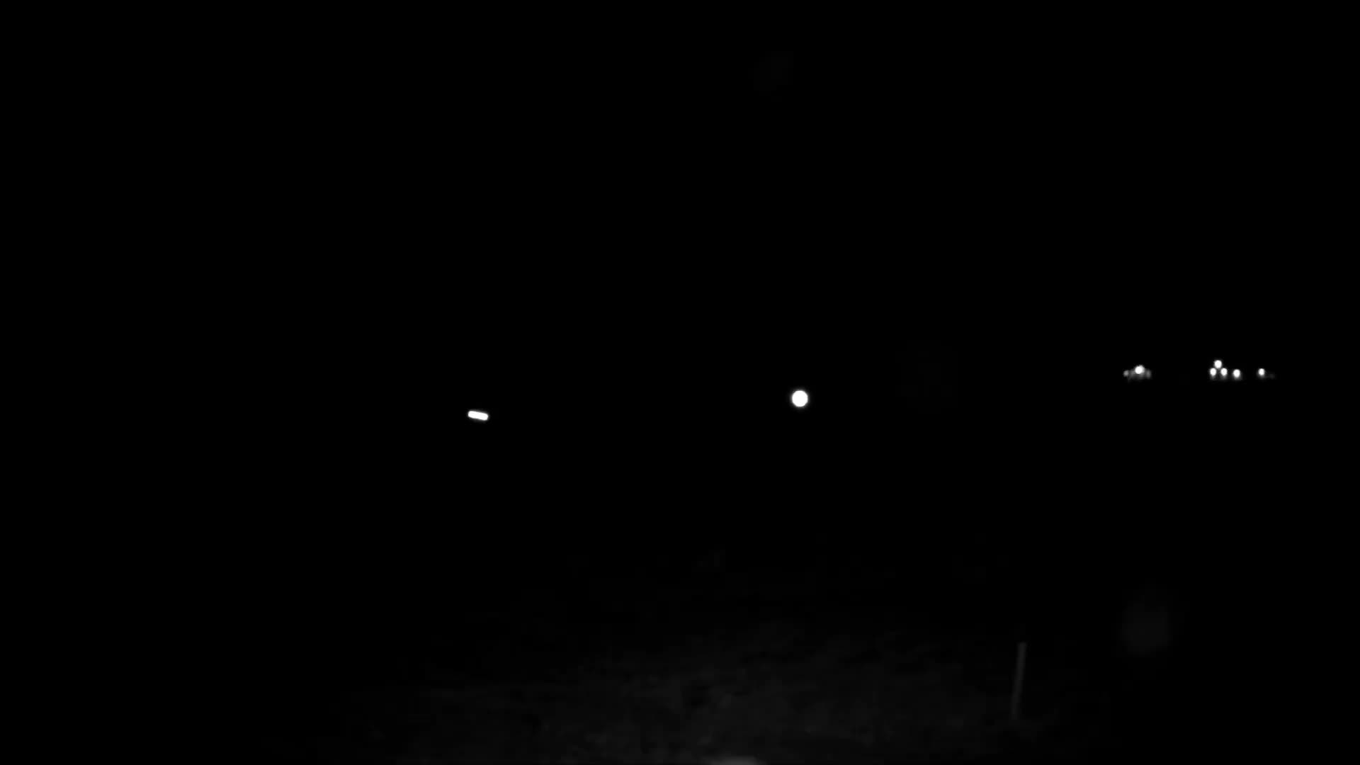 A clear, dark night scene features several distant, scattered white lights, likely from vehicles or structures, against an almost entirely black landscape with faint ground details visible at the bottom.