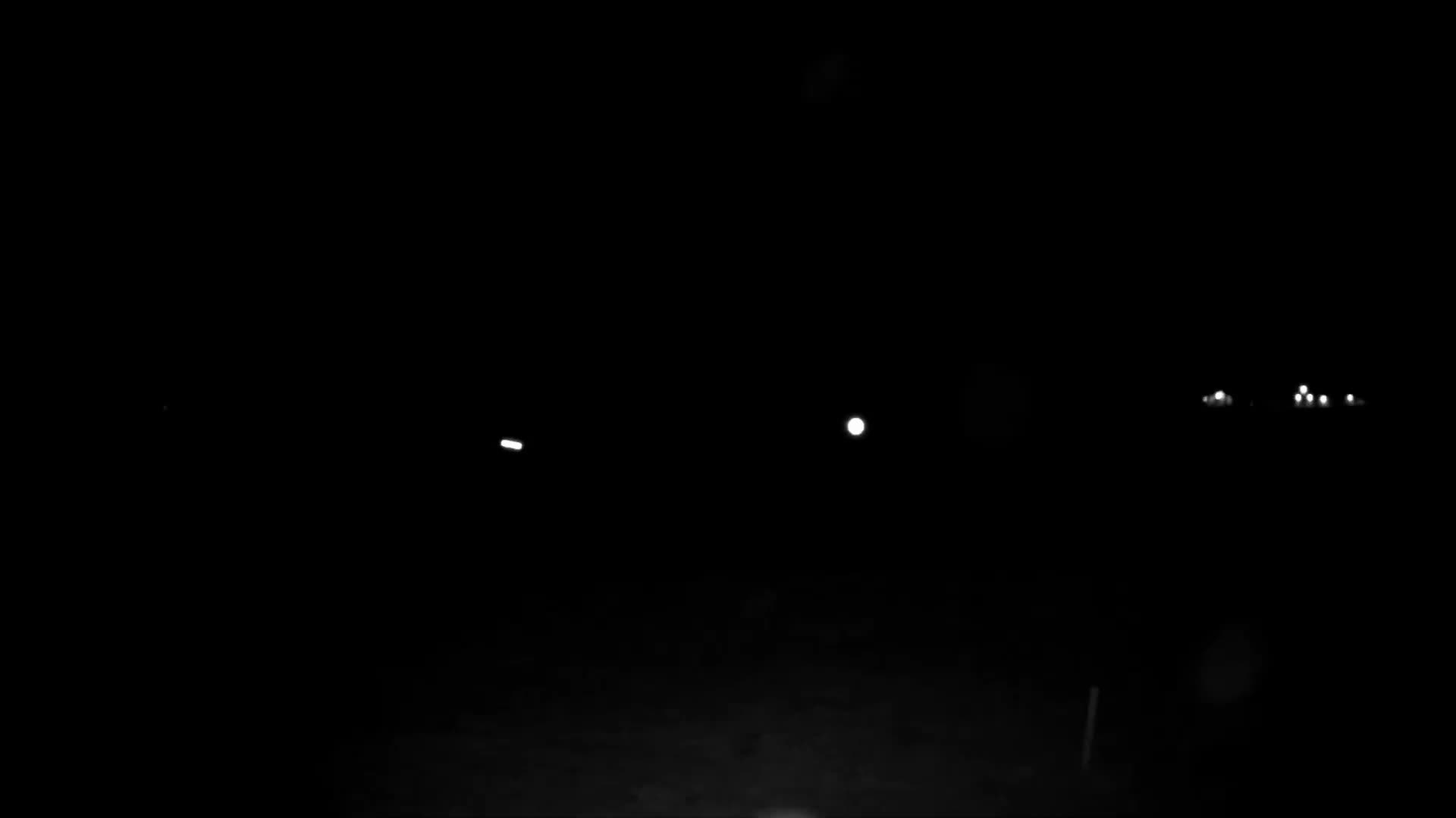 The image displays a very dark outdoor night scene, with sparse illumination from a singular elongated light on the left, a prominent round light in the center, and a cluster of smaller, blurry lights in the distance on the right.