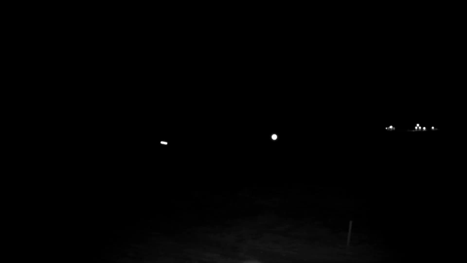The image displays a very dark outdoor night scene, with sparse illumination from a singular elongated light on the left, a prominent round light in the center, and a cluster of smaller, blurry lights in the distance on the right.