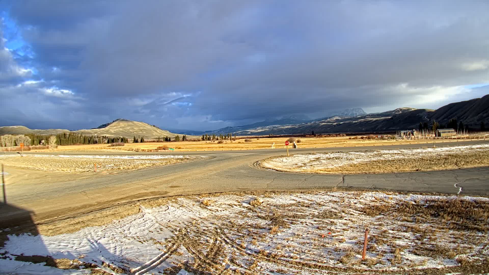 Under a dramatic, partly cloudy sky with dark storm clouds and intermittent sunlight, an expansive cold landscape features golden fields, scattered trees, distant mountains, and patches of snow on the foreground ground and intersecting dirt roads.