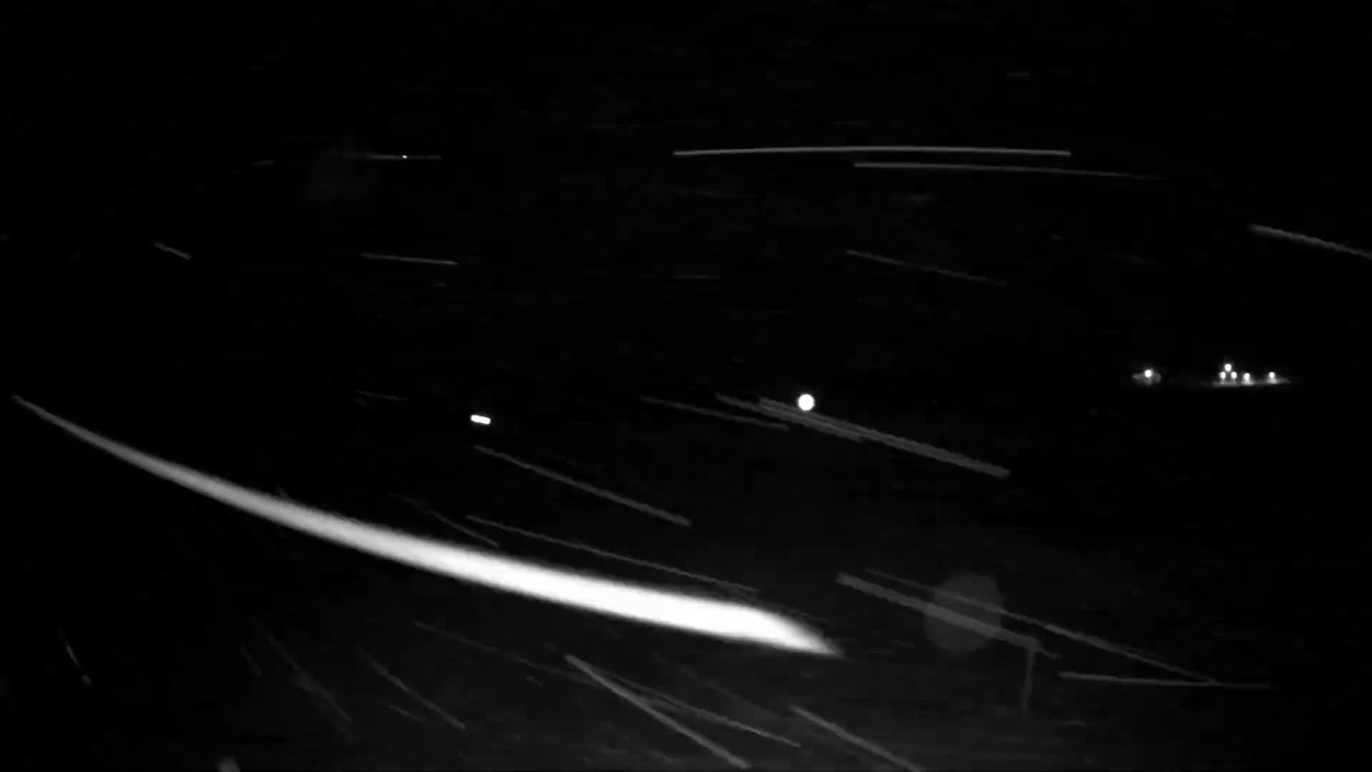 The image depicts a dark, nighttime outdoor scene with heavy snow falling, illuminated as numerous white streaks and dots, and a few faint distant lights visible on the right.