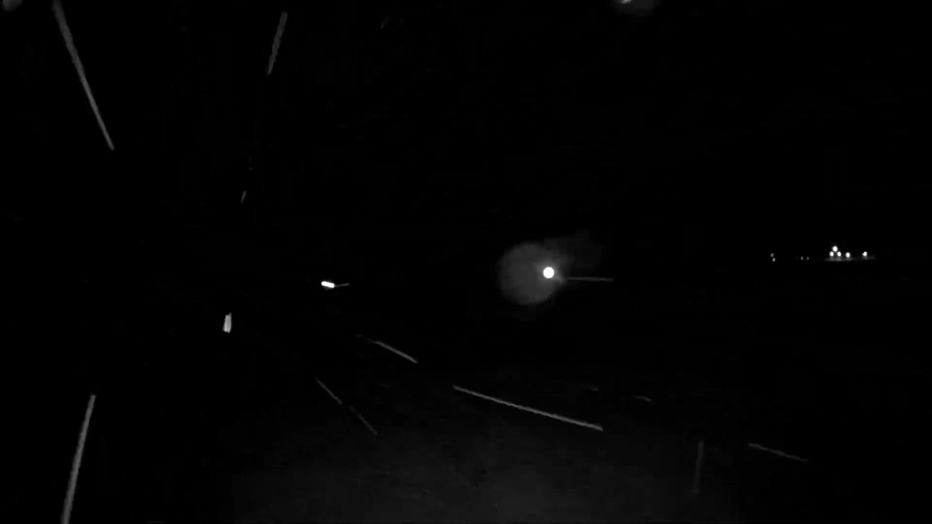The image depicts a dark, nighttime outdoor scene with heavy snow falling, illuminated as numerous white streaks and dots, and a few faint distant lights visible on the right.