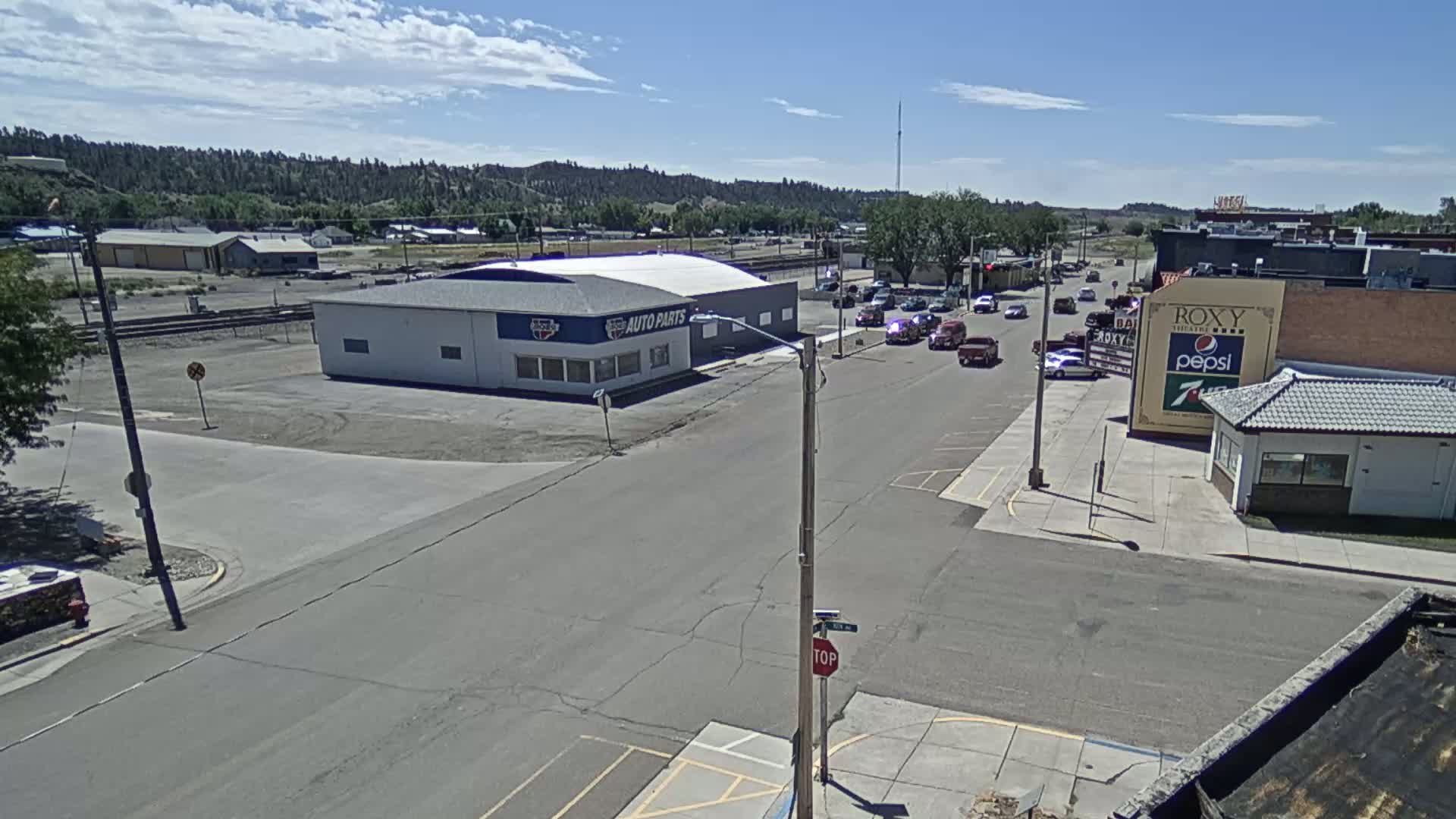 A high-angle view shows a small town street scene on a sunny day, with several vehicles traveling along the road past buildings including an auto parts store and a theater.