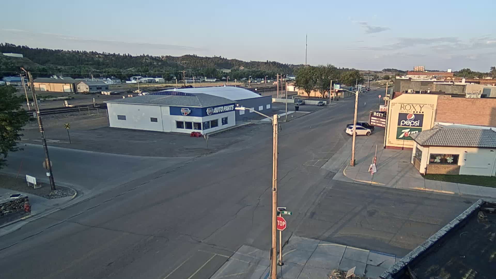A high-angle view shows a small town street scene on a sunny day, with several vehicles traveling along the road past buildings including an auto parts store and a theater.