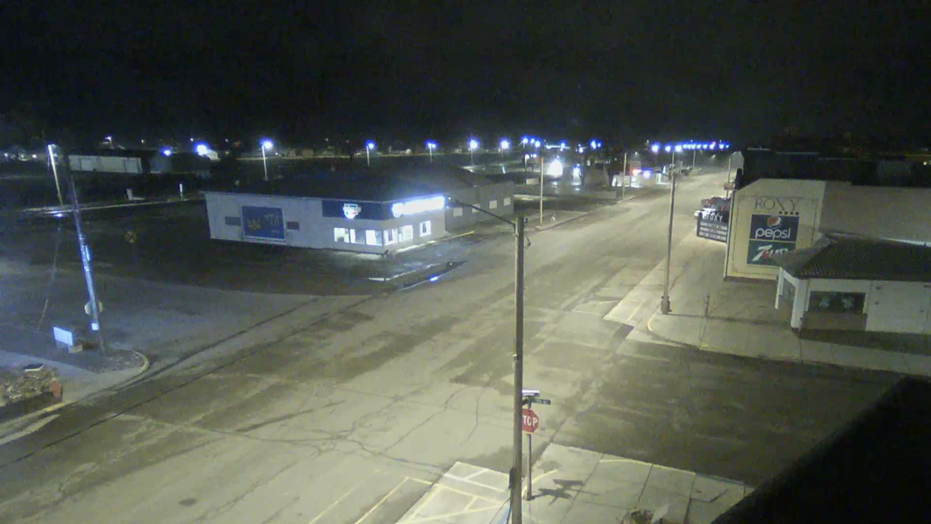 At night, a quiet urban street with wet asphalt is brightly illuminated by numerous streetlights, revealing commercial buildings under a clear sky.