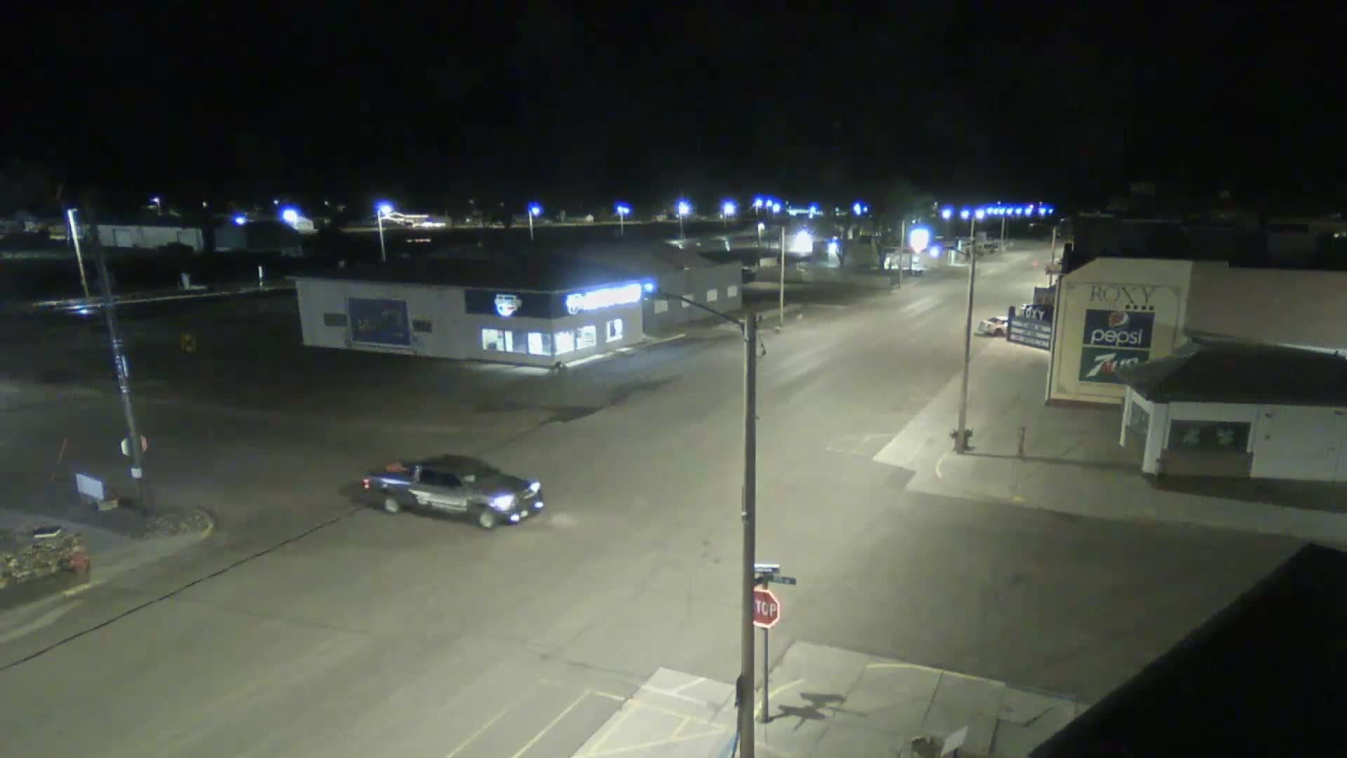 A clear night scene shows a dark pickup truck driving along a well-lit street lined with commercial buildings in what appears to be a small town.