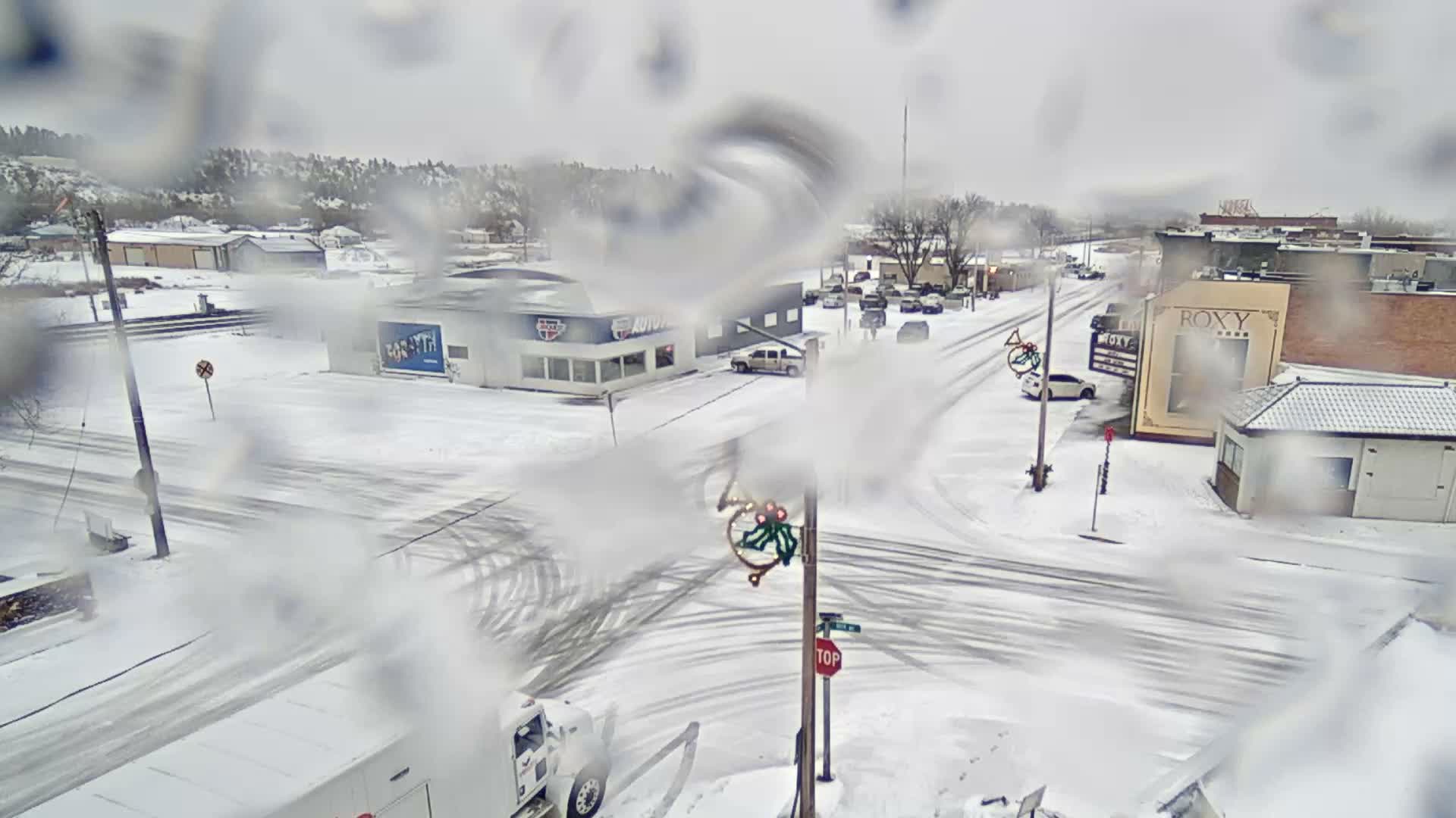 A snow-covered town scene, with buildings, roads, and parked vehicles blanketed in white, is visible through a moisture-streaked lens under snowy, overcast conditions.