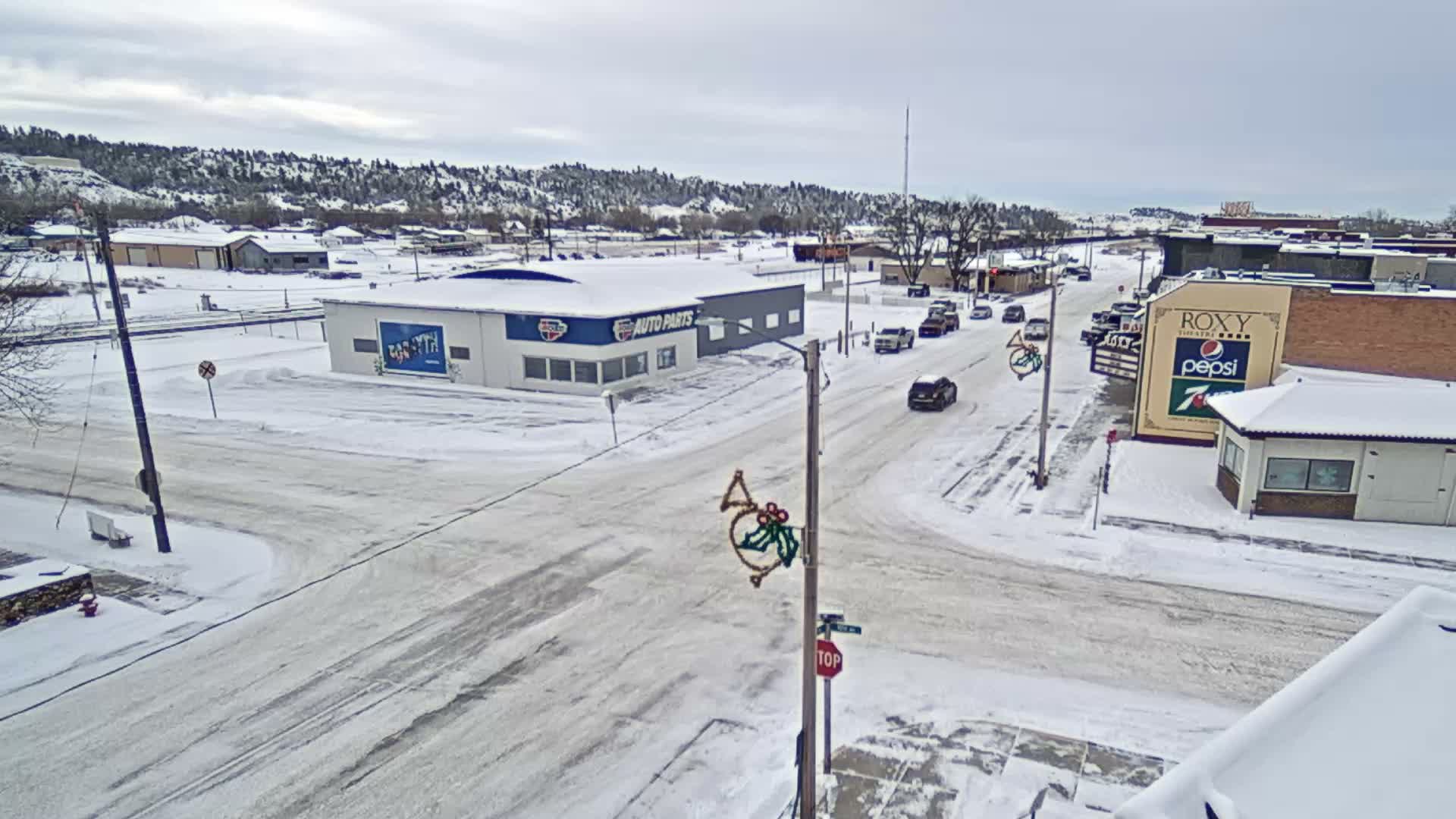 An elevated view on a clear night shows a quiet street illuminated by streetlights, with buildings on both sides, one adorned with blue festive lights, and a holiday figure mounted on a utility pole.