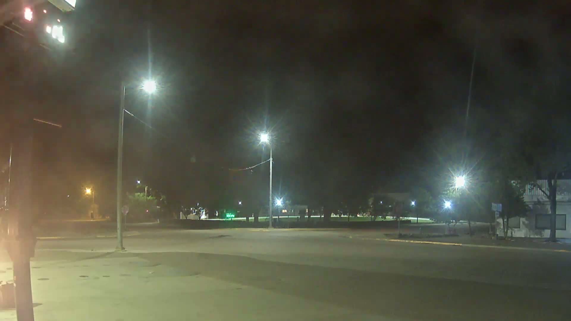 A nighttime view of an empty intersection with streetlights illuminating a small park and a building in the background.