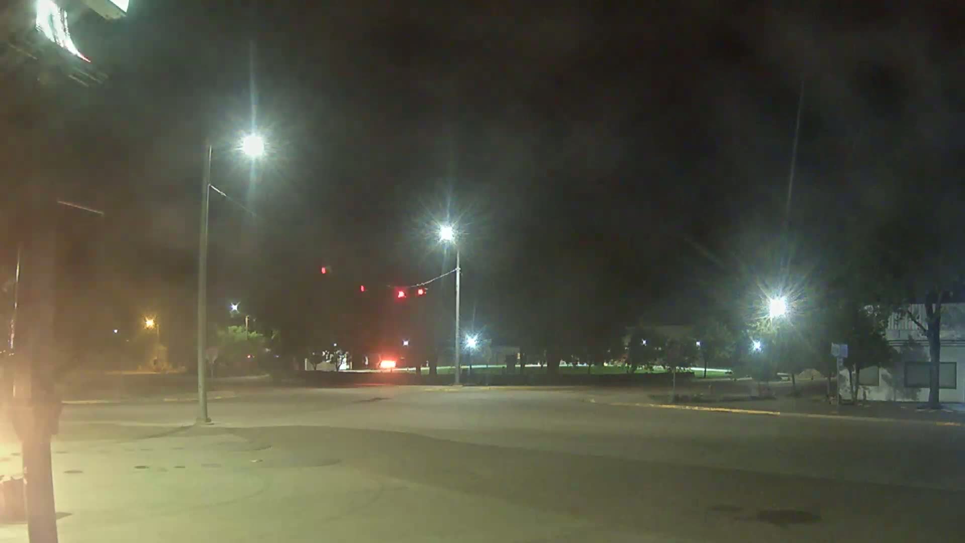 A nighttime view of an empty intersection with streetlights illuminating a small park and a building in the background.