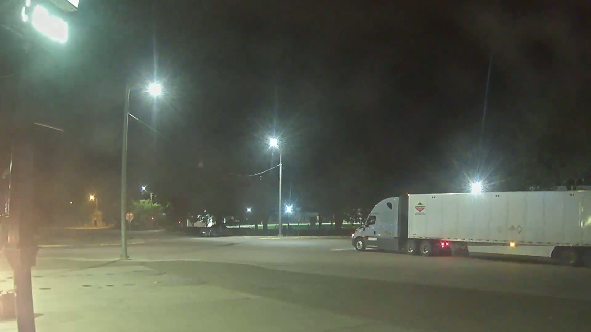 At night, a large semi-truck with a white trailer is parked in a dimly lit lot, with several streetlights illuminating the scene.