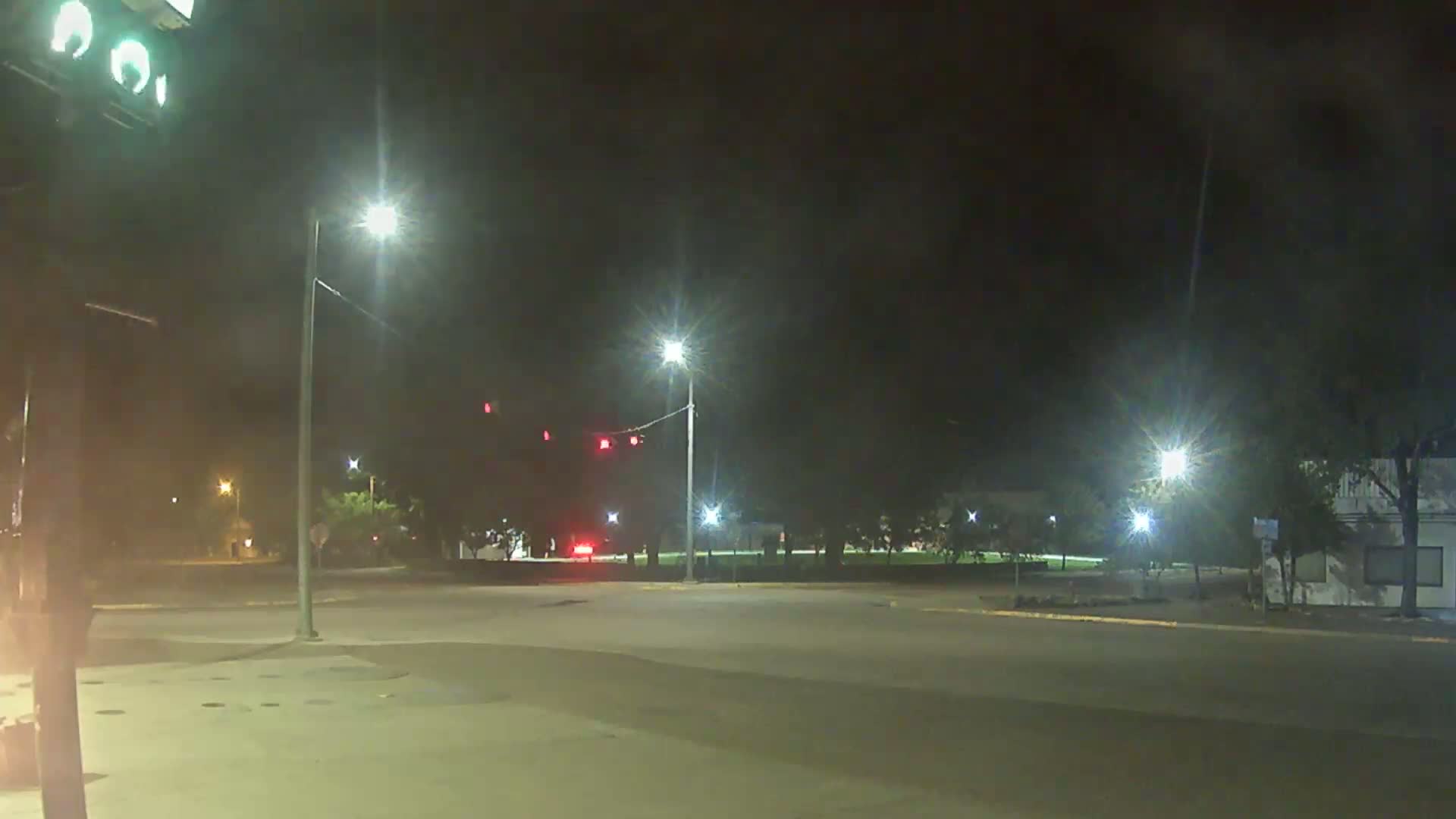 A nighttime view of a dimly lit intersection with streetlights, some trees, and a few vehicles visible in the distance under a hazy sky.