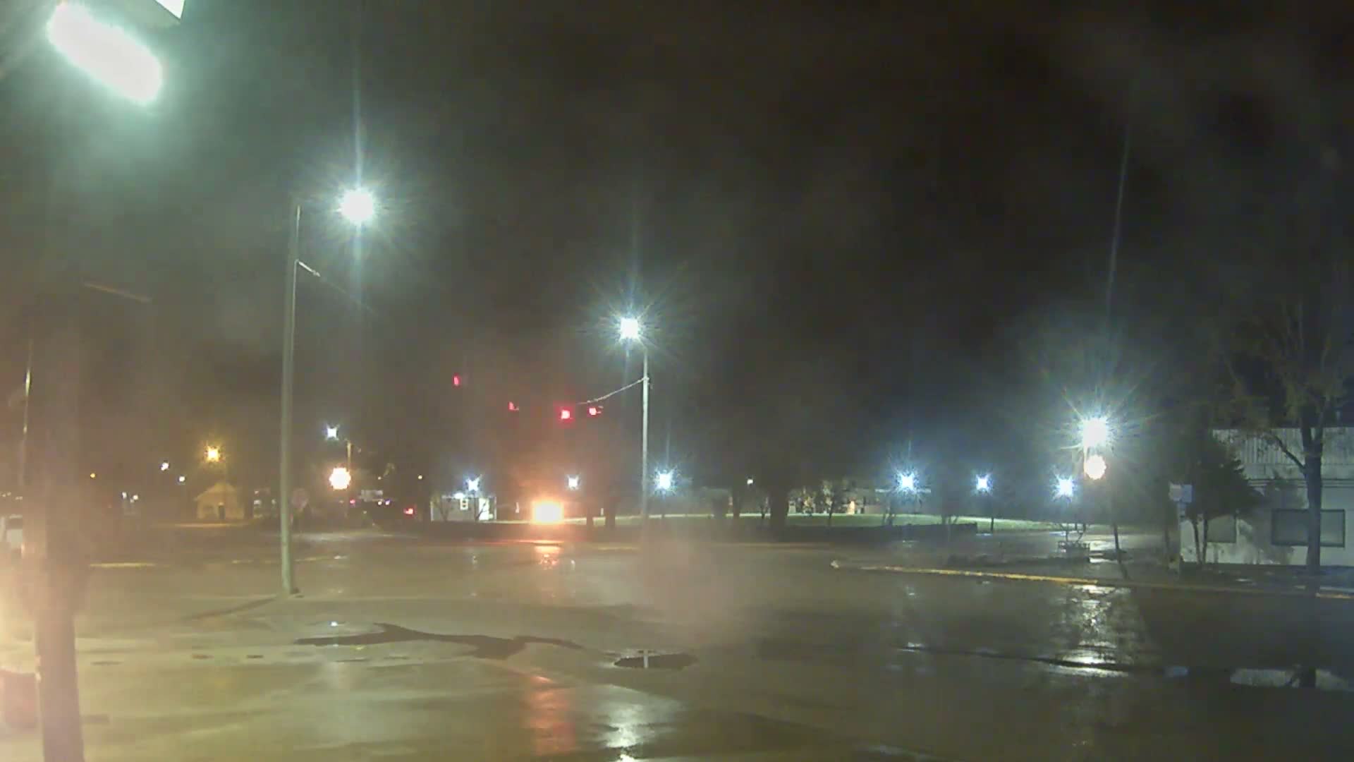 A street intersection appears brightly lit by numerous streetlights and traffic signals on a dark, wet night, with reflections on the rain-slicked pavement and some steam rising from a grate.