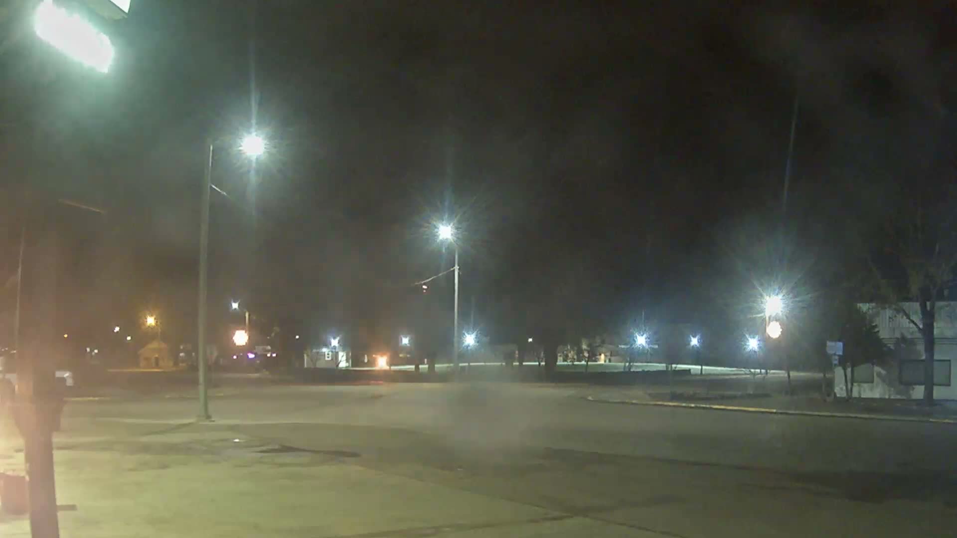 A clear night illuminates an empty urban intersection with numerous bright streetlights and distant building lights under a dark sky.