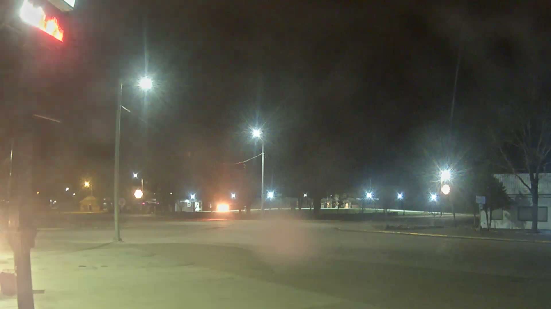 A hazy night scene captures a fire burning on a building's roof on the left, with multiple bright streetlights illuminating a street and bare trees under a dark sky.