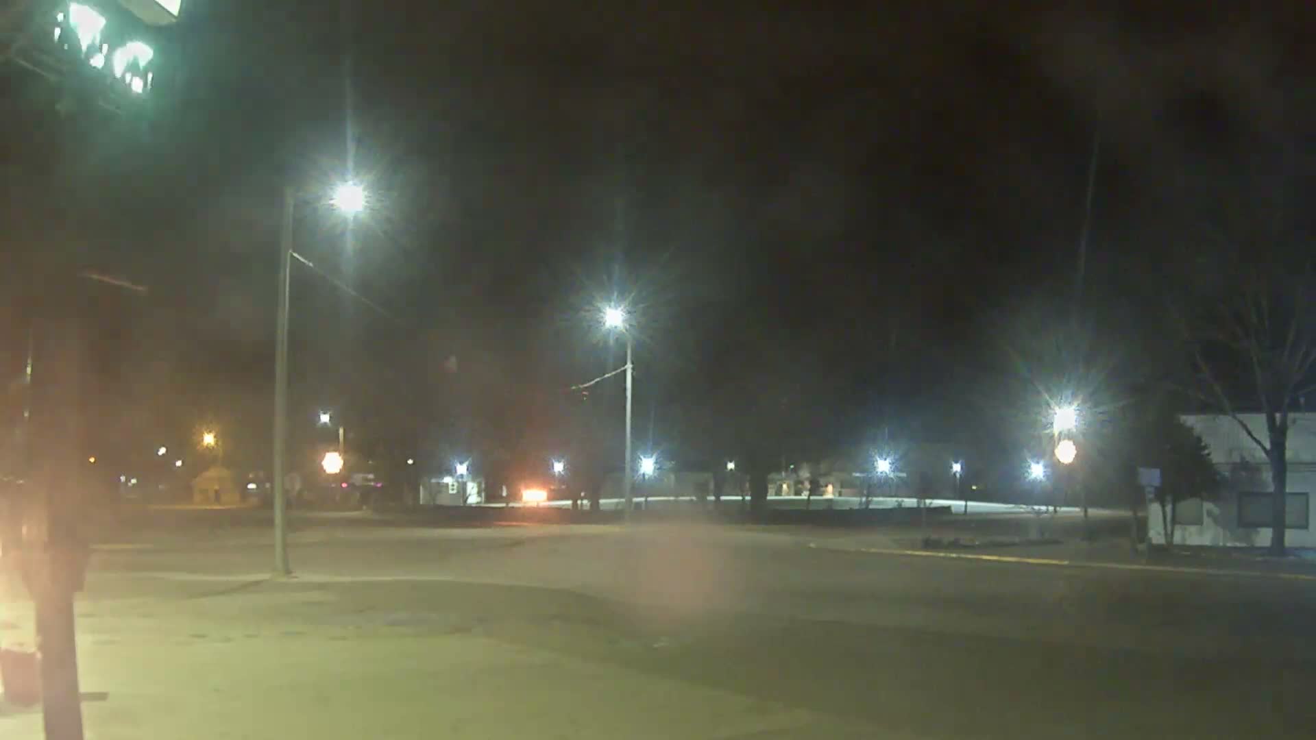 A hazy night scene captures a fire burning on a building's roof on the left, with multiple bright streetlights illuminating a street and bare trees under a dark sky.