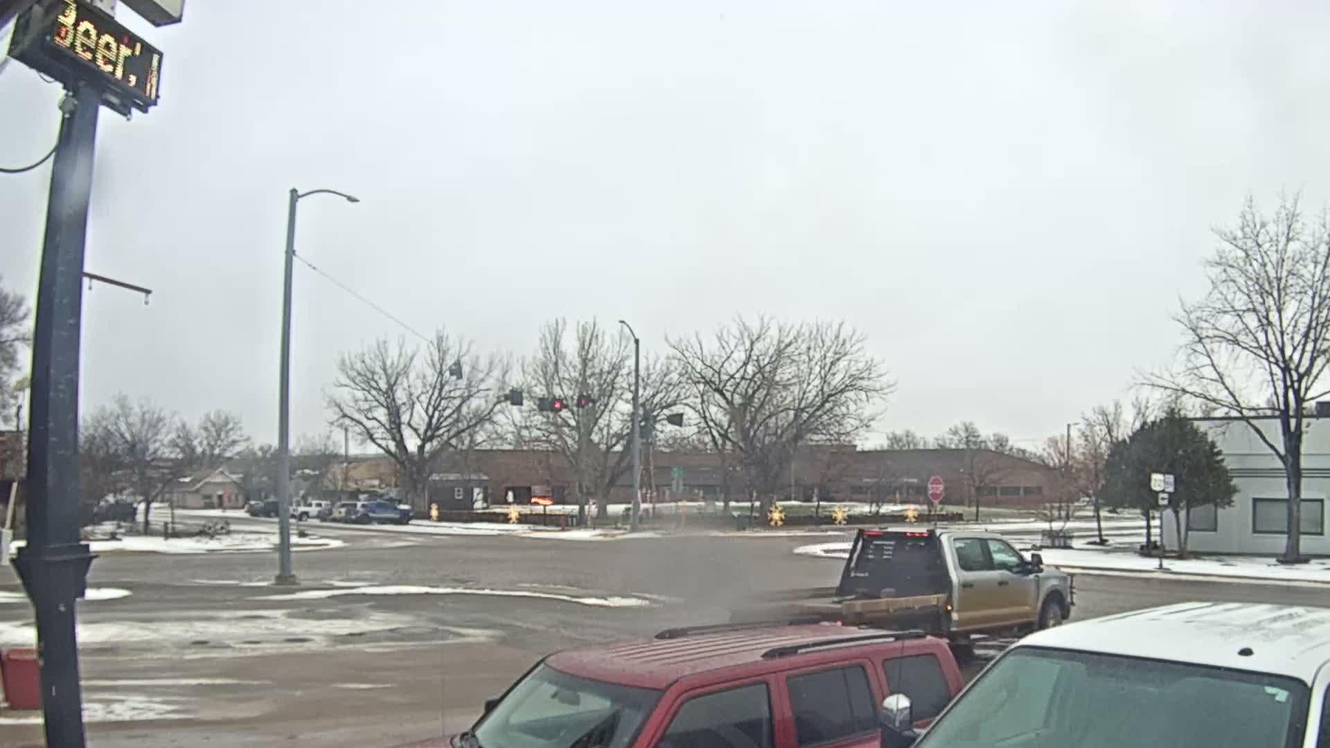 A wintry, overcast day reveals an intersection with bare trees and slushy roads, featuring vehicles like a red SUV in the foreground and a silver flatbed truck.