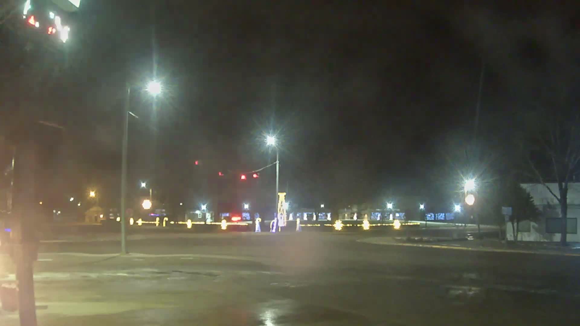 A wet and hazy night reveals a town square or intersection illuminated by streetlights, red traffic signals, and numerous festive holiday lights decorating the ground and buildings.