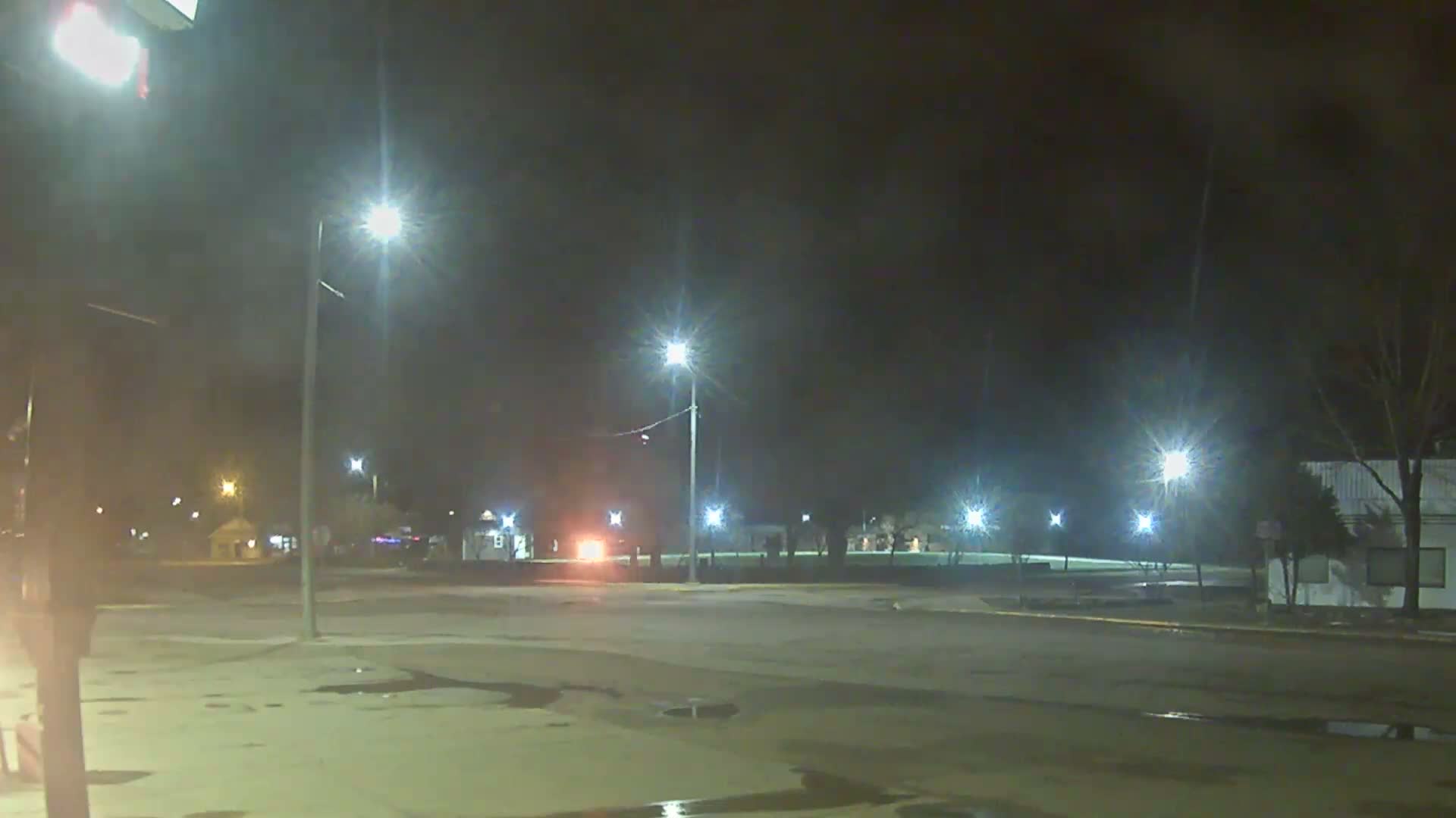 A wet and hazy night reveals a town square or intersection illuminated by streetlights, red traffic signals, and numerous festive holiday lights decorating the ground and buildings.