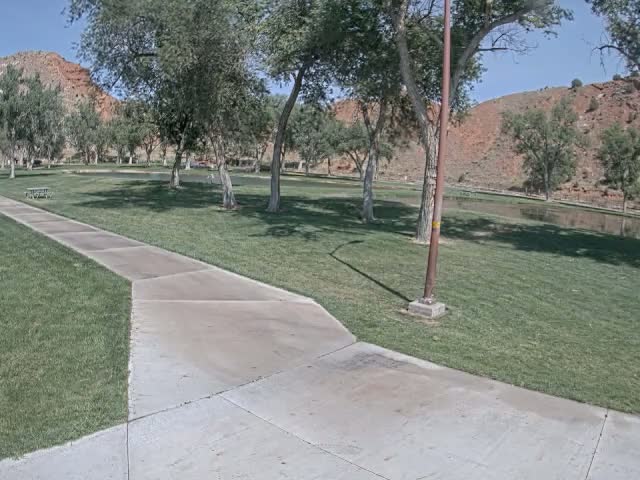 A paved pathway winds through a grassy park with several trees under a sunny sky, near a calm body of water and a red rock hillside in the background.