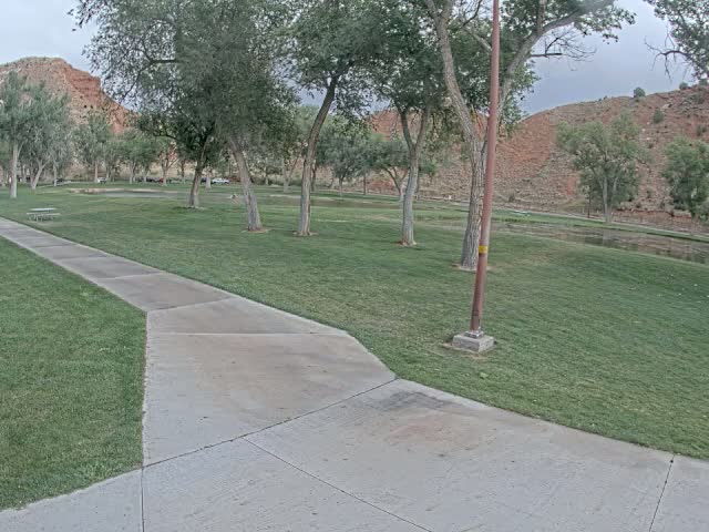 A paved pathway winds through a grassy park with several trees under a sunny sky, near a calm body of water and a red rock hillside in the background.