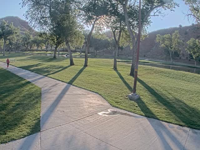 A paved pathway winds through a grassy park with several trees under a sunny sky, near a calm body of water and a red rock hillside in the background.