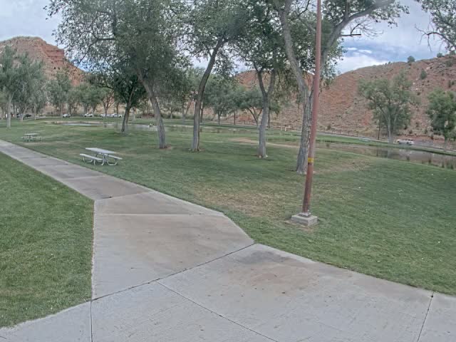 A paved pathway leads through a grassy park with picnic tables, several trees, and a small pond, all set against a backdrop of reddish-brown hills under a partly cloudy sky.