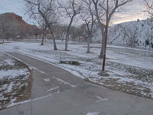A wintry outdoor scene shows a paved path cutting through snow-dusted grass and bare trees, with a red mountain on the left and a snow-covered hill across a canal on the right, all under a cloudy sky.