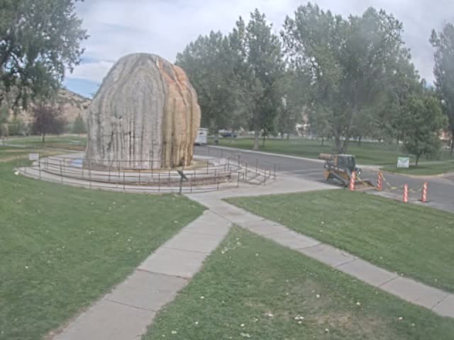 A large, light-colored rock formation, possibly a petrified geyser, sits in a park-like setting under an overcast sky, with some light construction equipment visible nearby.