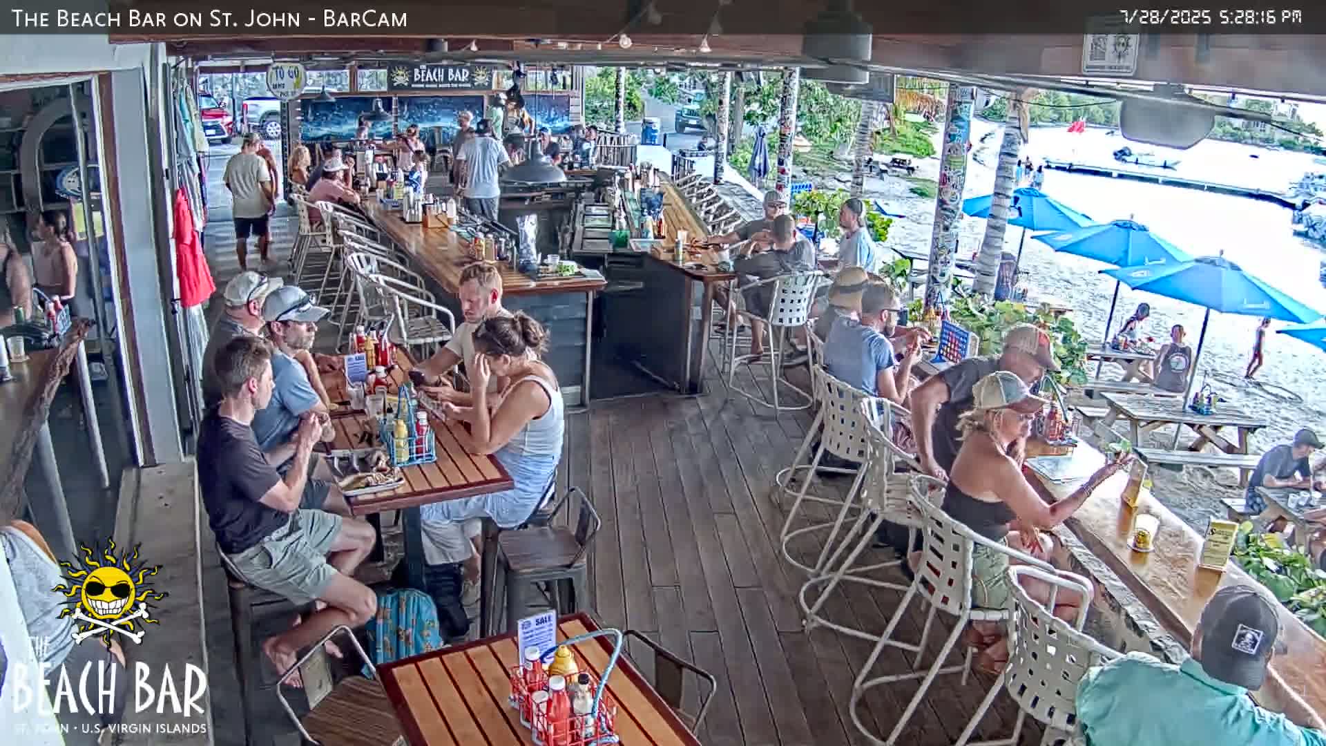 An outdoor beach bar with patrons seated at various tables under blue umbrellas on a sunny day.