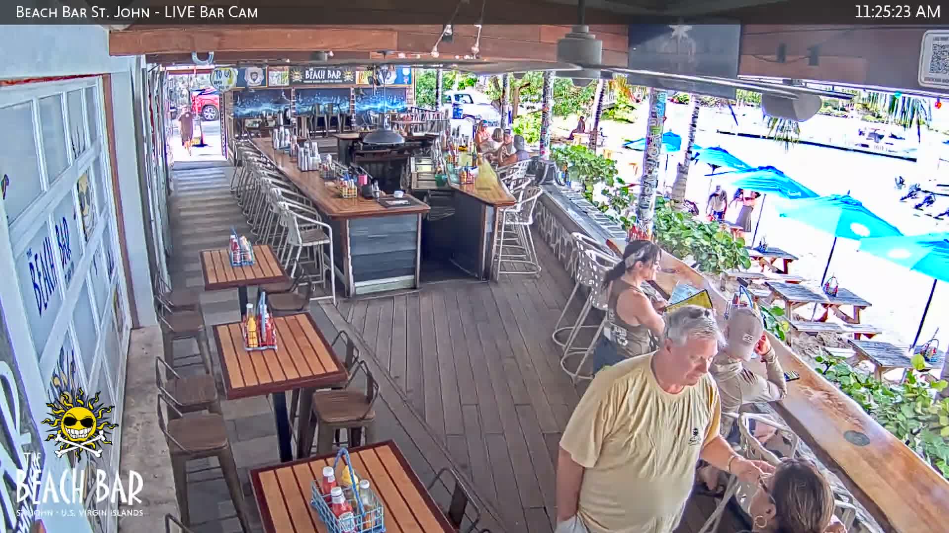 Patrons are seen enjoying a clear, sunny day at an open-air bar and restaurant, which features a long counter, several dining tables, and an outdoor seating area with blue umbrellas overlooking what appears to be a waterfront.