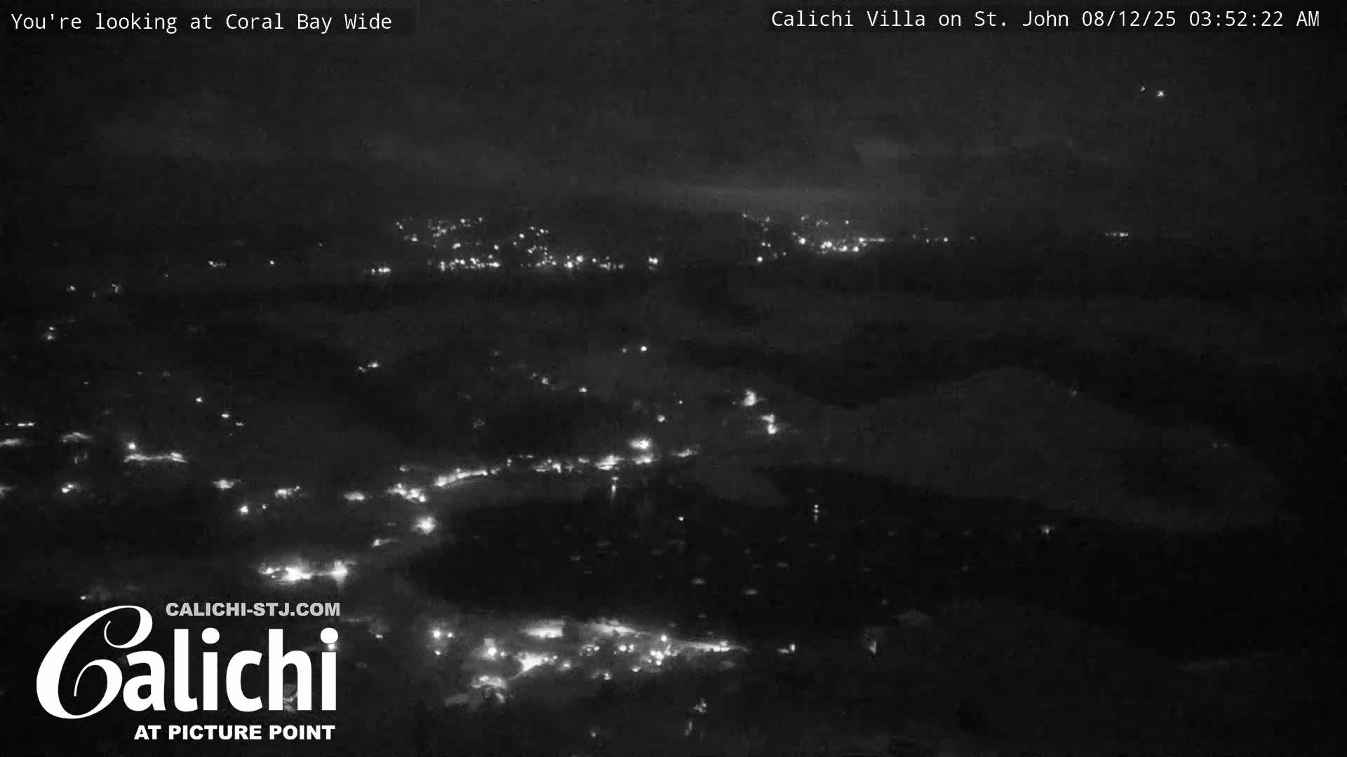 A nighttime, wide shot shows a dark landscape with scattered lights from a town below, under a mostly cloudy sky.