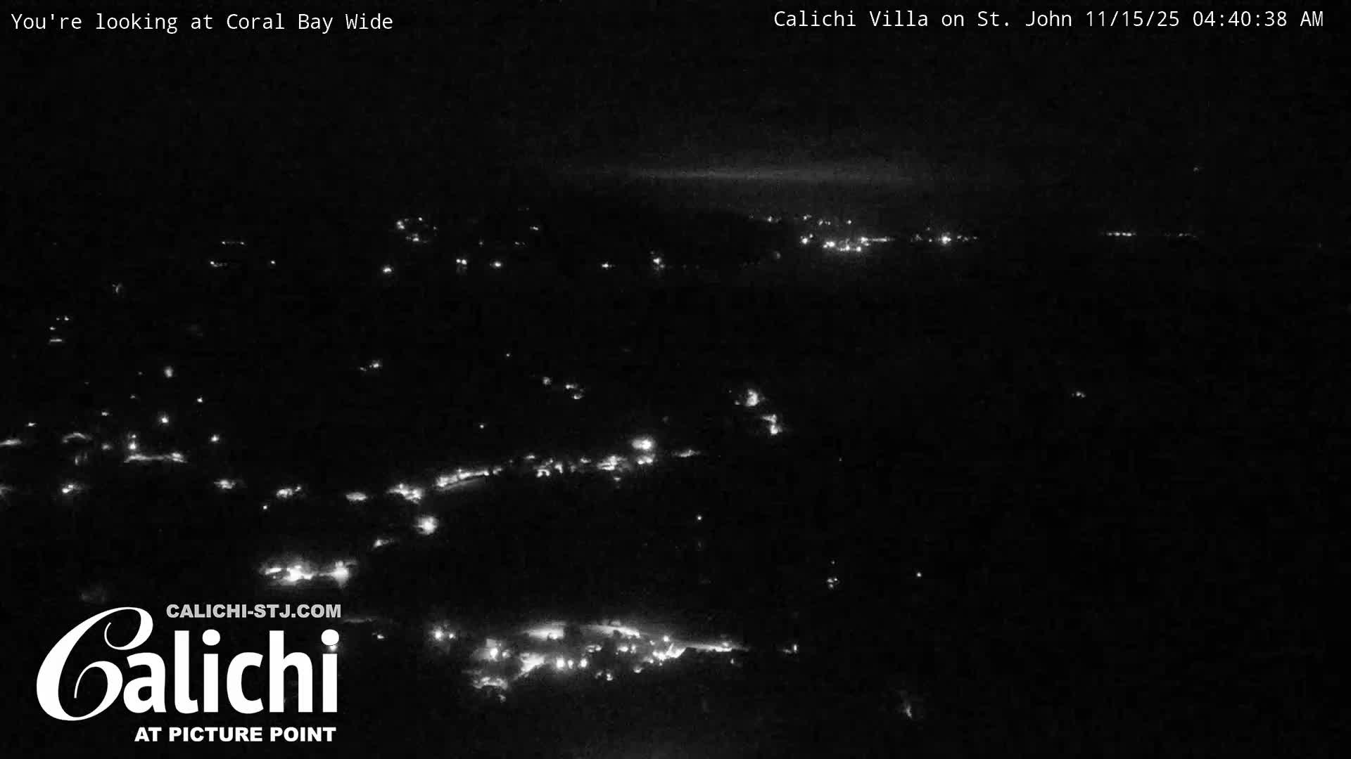 A dark, wide-angle night view reveals numerous scattered lights illuminating a coastal or island settlement under clear skies.