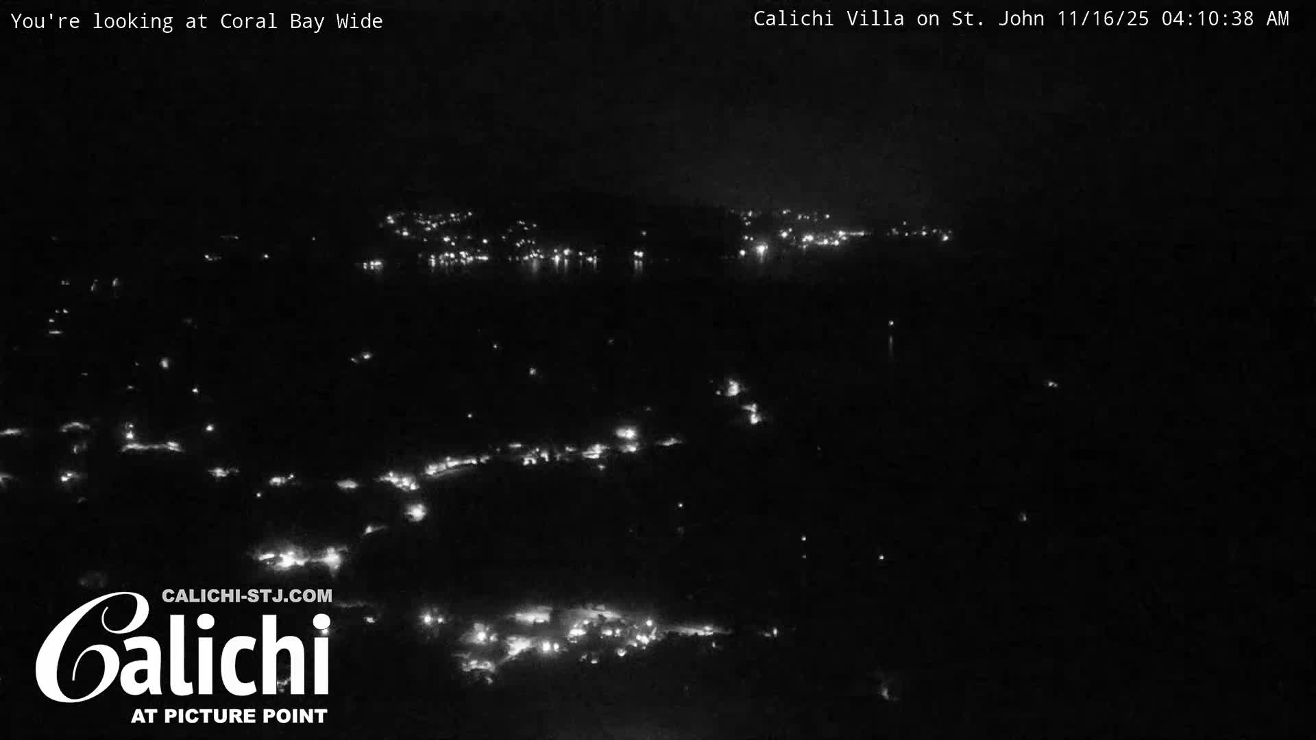 A dark, clear night scene reveals scattered artificial lights from a coastal settlement illuminating a shoreline across a dark expanse of water.