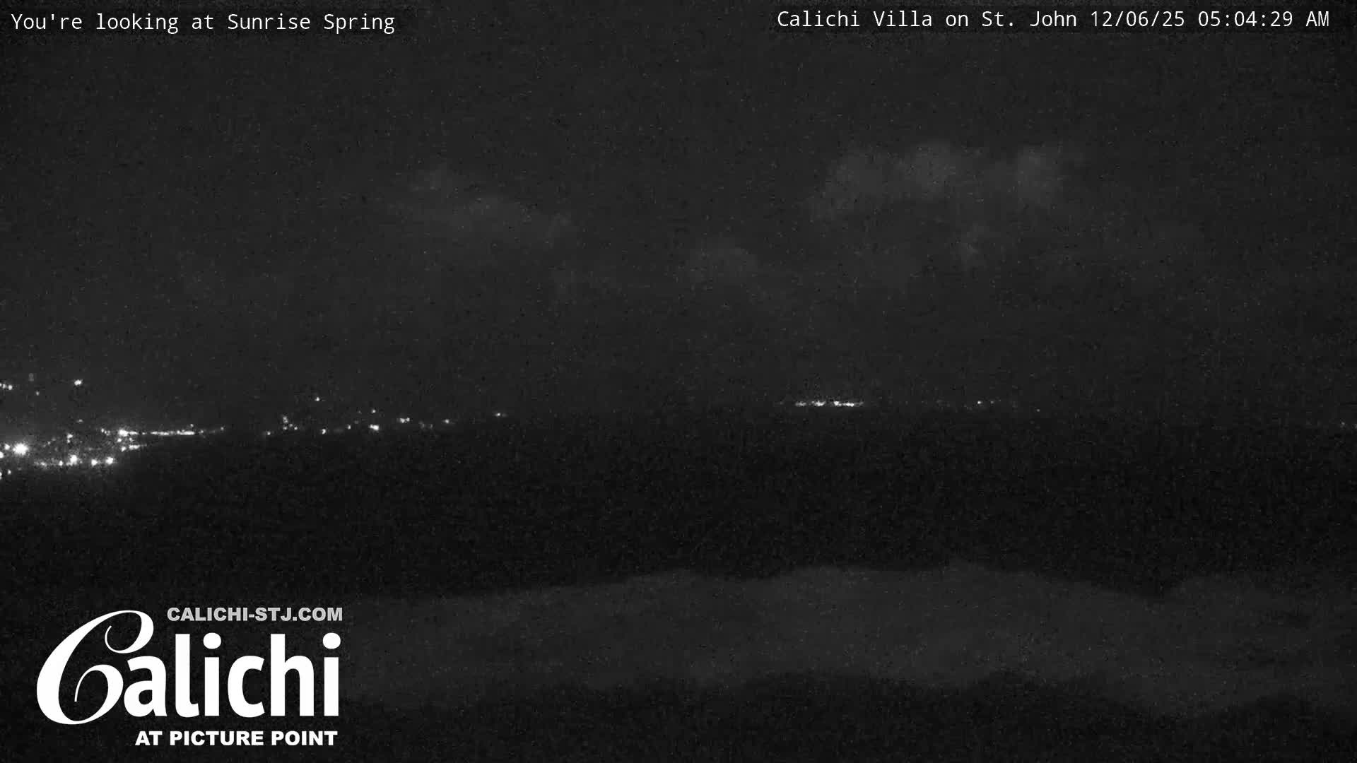 A dark, nighttime outdoor scene displays scattered clusters of distant lights across a landscape, beneath a partly cloudy sky with a faint, low-lying mist or fog.