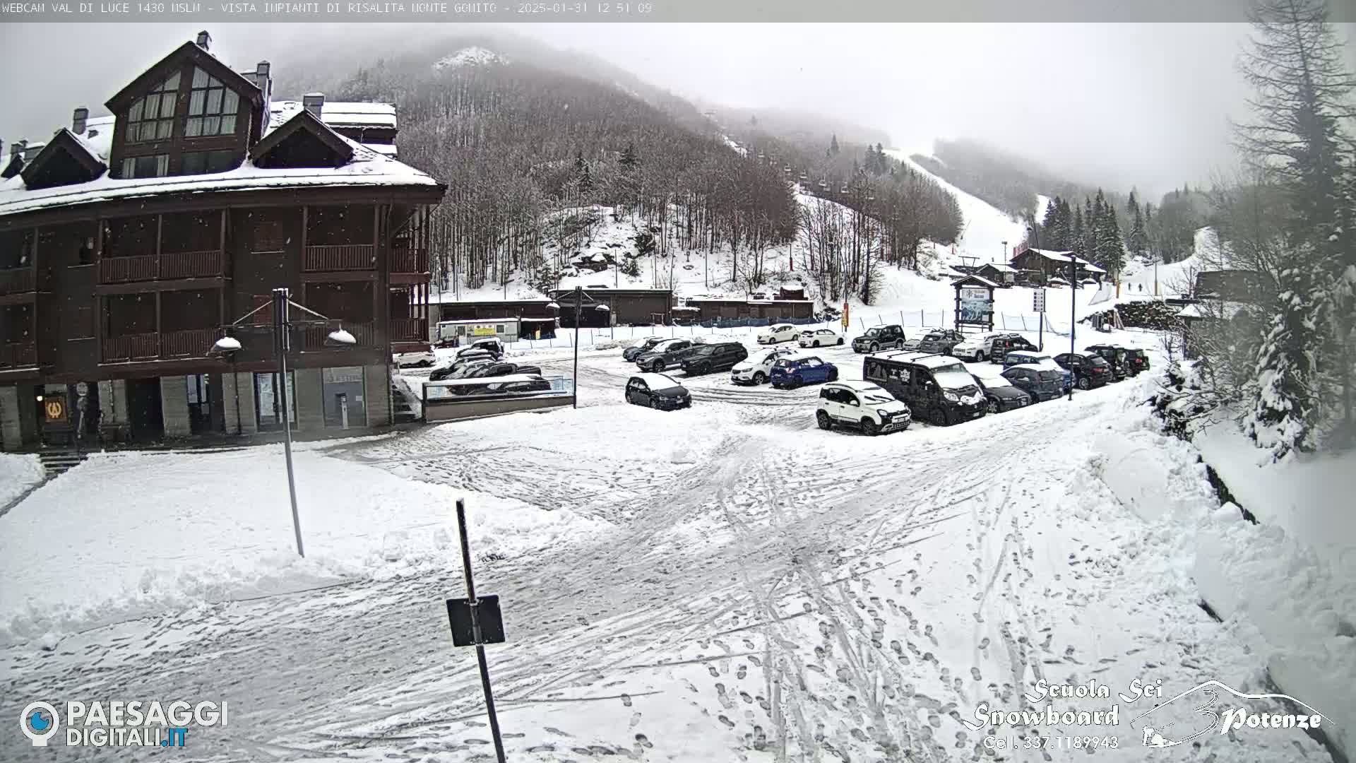 A snow-covered parking lot with cars and a multi-story building is situated in front of a snow-covered mountain slope with a ski lift visible in the background under overcast conditions.