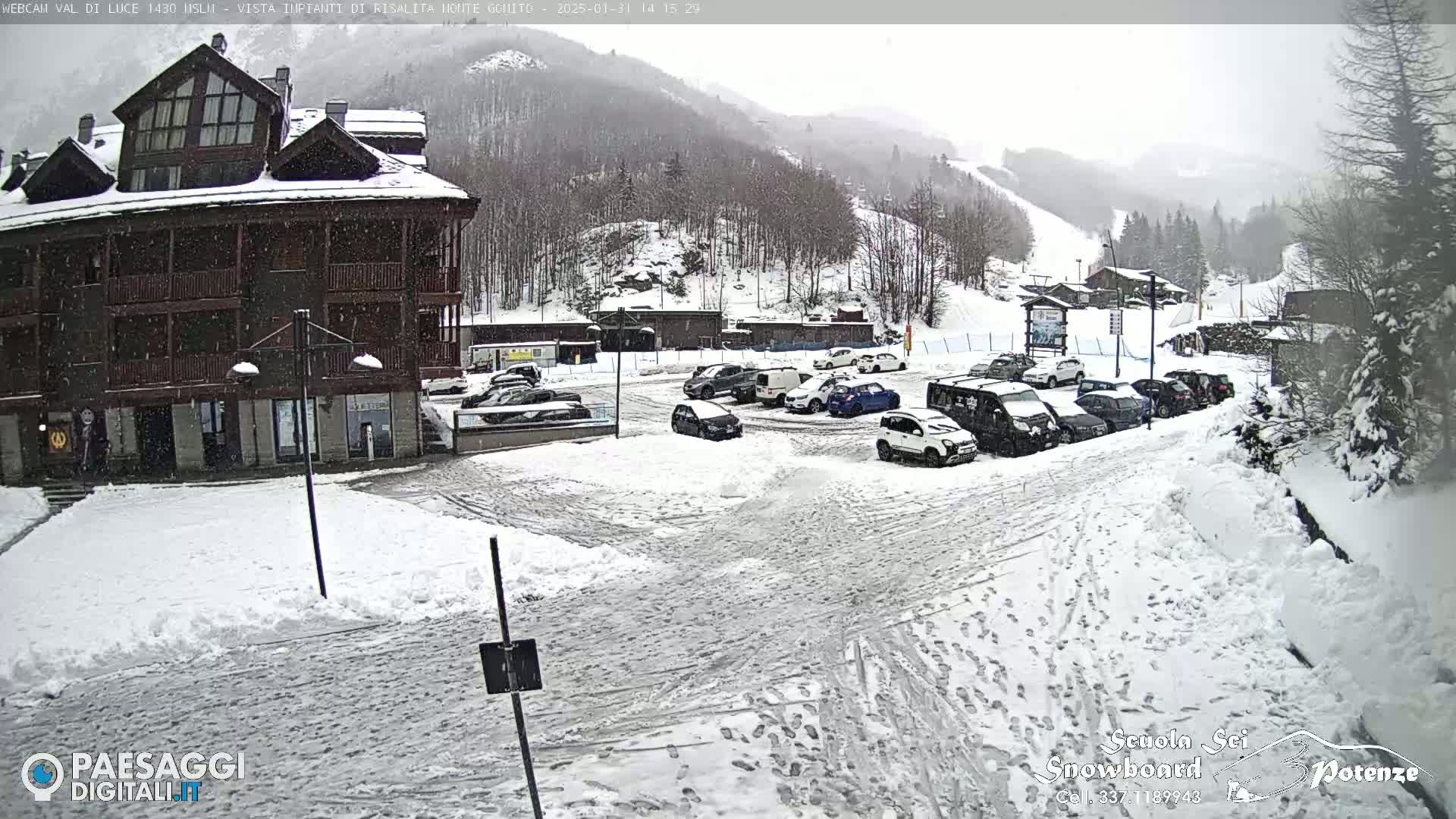 A snow-covered parking lot with cars in front of a multi-story building, situated amidst snowy mountains under a light snowfall.