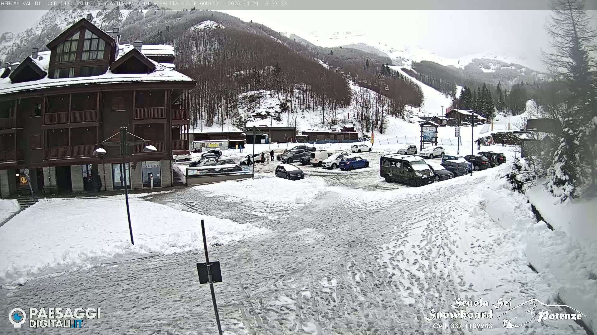 A snow-covered parking lot with several cars and a multi-story building is situated in front of a snow-dusted mountainside.