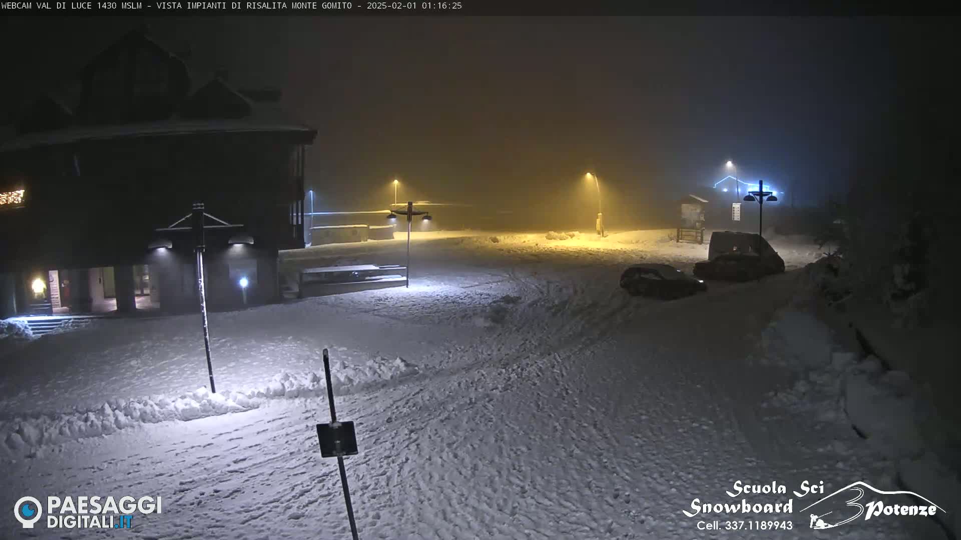 A snow-covered parking lot at night is dimly lit by streetlights in foggy conditions, with a building and a few vehicles visible.